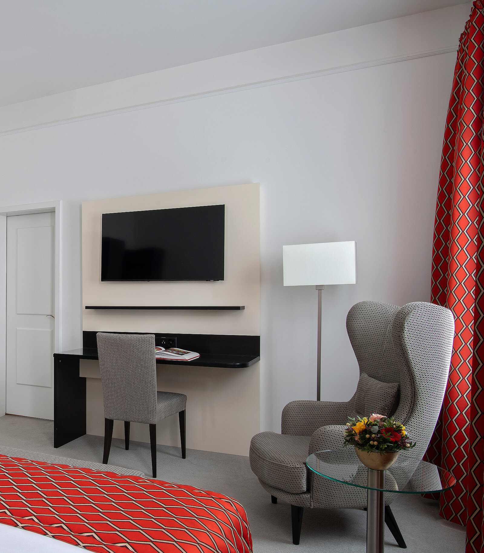 Ein stilvoll eingerichtetes Hotelzimmer mit einem großen Fernseher an der Wand, einem Schreibtisch mit Stuhl darunter und einem Sessel neben einem kleinen runden Tisch. Rote Vorhänge mit geometrischem Muster hängen an der Seite.

