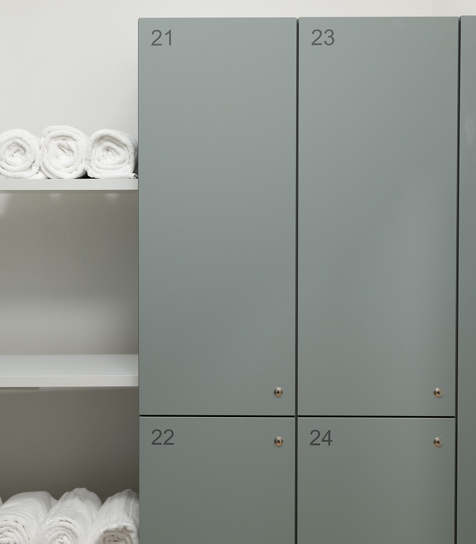 Graue Spinde mit den Nummern 21, 22, 23 und 24 sind zu sehen. Links davon befinden sich gestapelte, sauber gefaltete Handtücher auf Regalen.