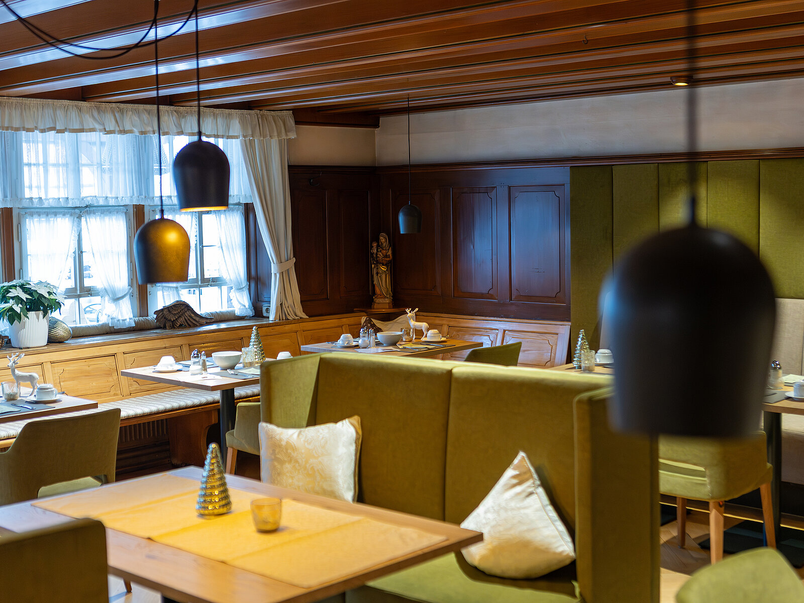 Gemütliches Restaurant mit Holztäfelung und mehreren Holztischen, die mit gelben Tischsets gedeckt sind. Grüne Polsterbänke und -stühle bieten Sitzgelegenheiten. Warmes Licht von Hängelampen schafft eine einladende Atmosphäre. Fensterscheiben mit Vorhängen lassen Tageslicht herein.