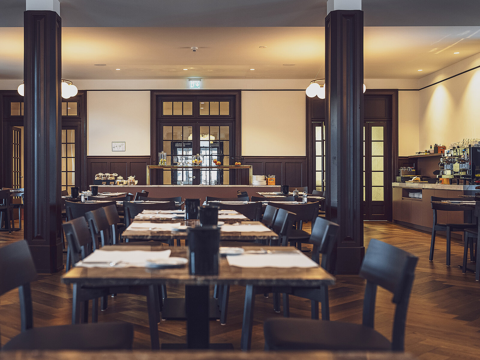 Ein elegantes Restaurant mit gedeckten Tischen, dunklen Holzstühlen und gedämpftem Licht. Im Hintergrund sind Getränkestationen und Retro-Elemente zu sehen. Die Atmosphäre wirkt einladend und stilvoll.