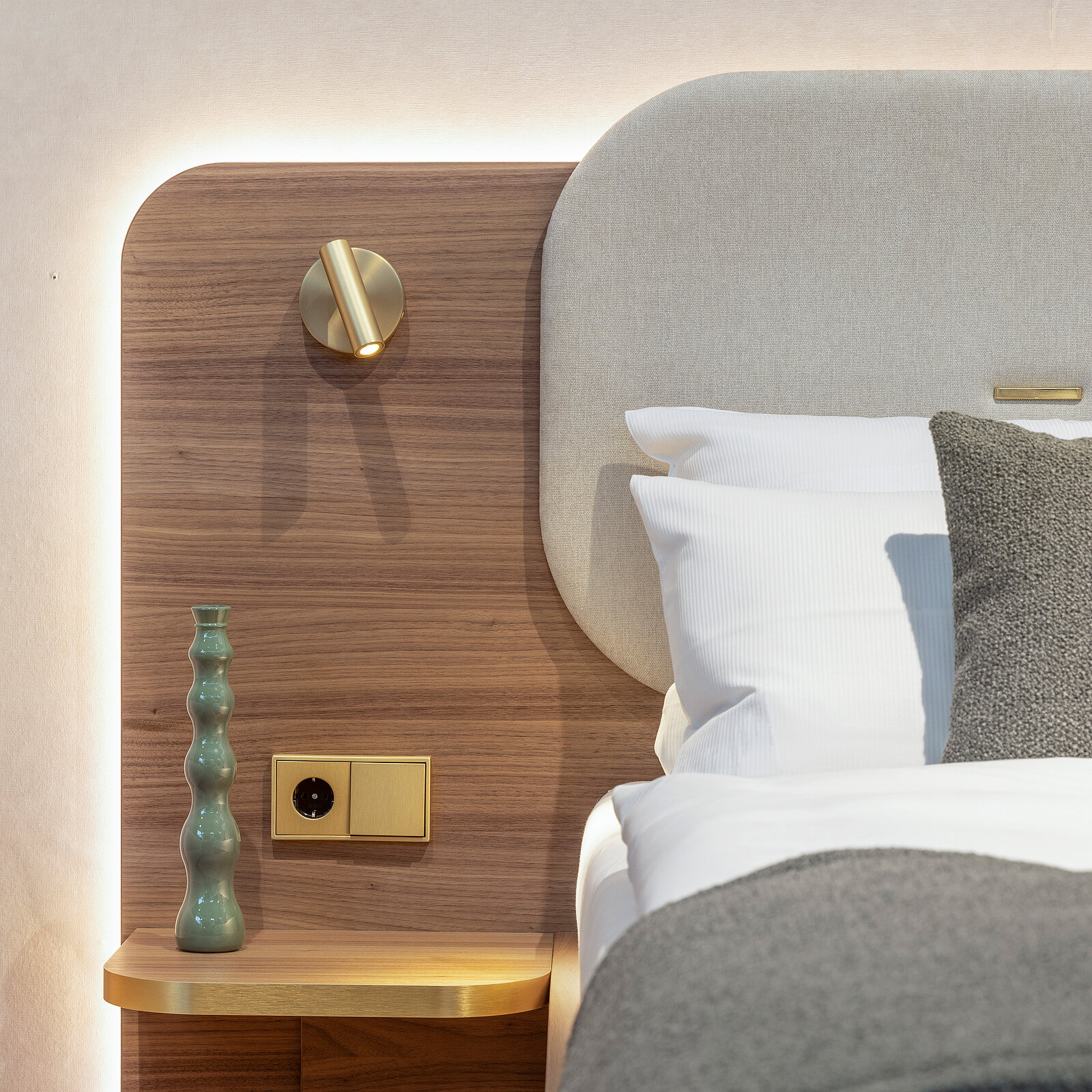 A modern bedroom features a stylish headboard with soft lighting. A textured gray blanket covers the bed, and decorative elements include a wooden nightstand with a green decorative vase and a light switch. Warm wood accents create a cozy atmosphere.
