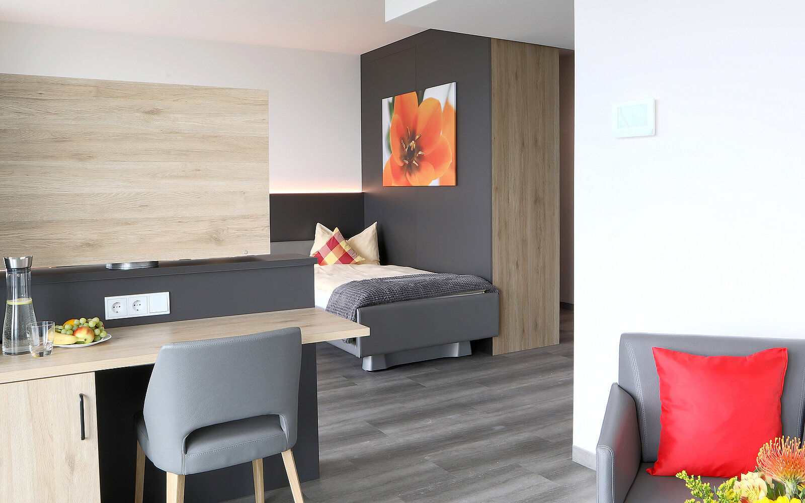 Das Bild zeigt eine moderne Hotelzimmer-Inneneinrichtung mit einem Essbereich, einem Bett und einem gemütlichen Sessel. Die Wände sind in neutralen Farben gehalten, und ein großes Kunstwerk einer Blume hängt über dem Bett. Der Raum wirkt hell und einladend.