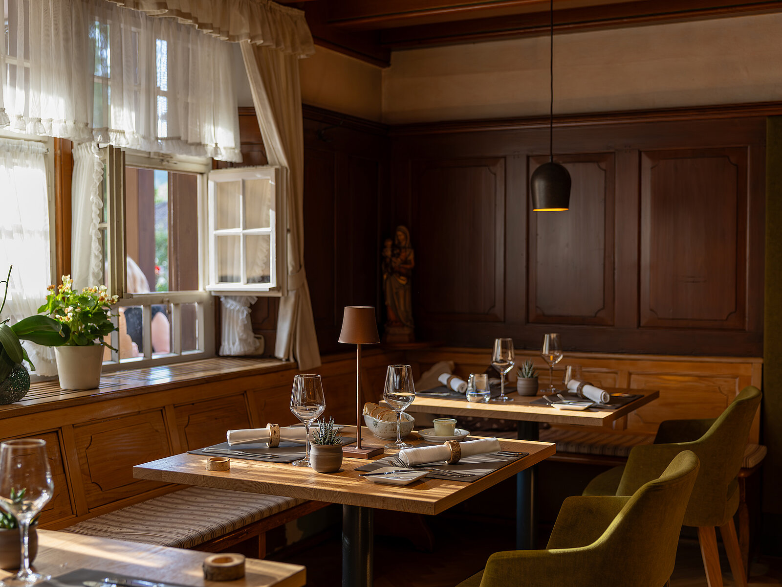 Gemütlich eingerichtetes Restaurant mit Holztischen, gedeckt mit Gläsern, Tellern und Stoffservietten. Warme Beleuchtung schafft eine einladende Atmosphäre. Pflanze auf dem Fensterbrett und Holzwandverkleidung im Hintergrund.