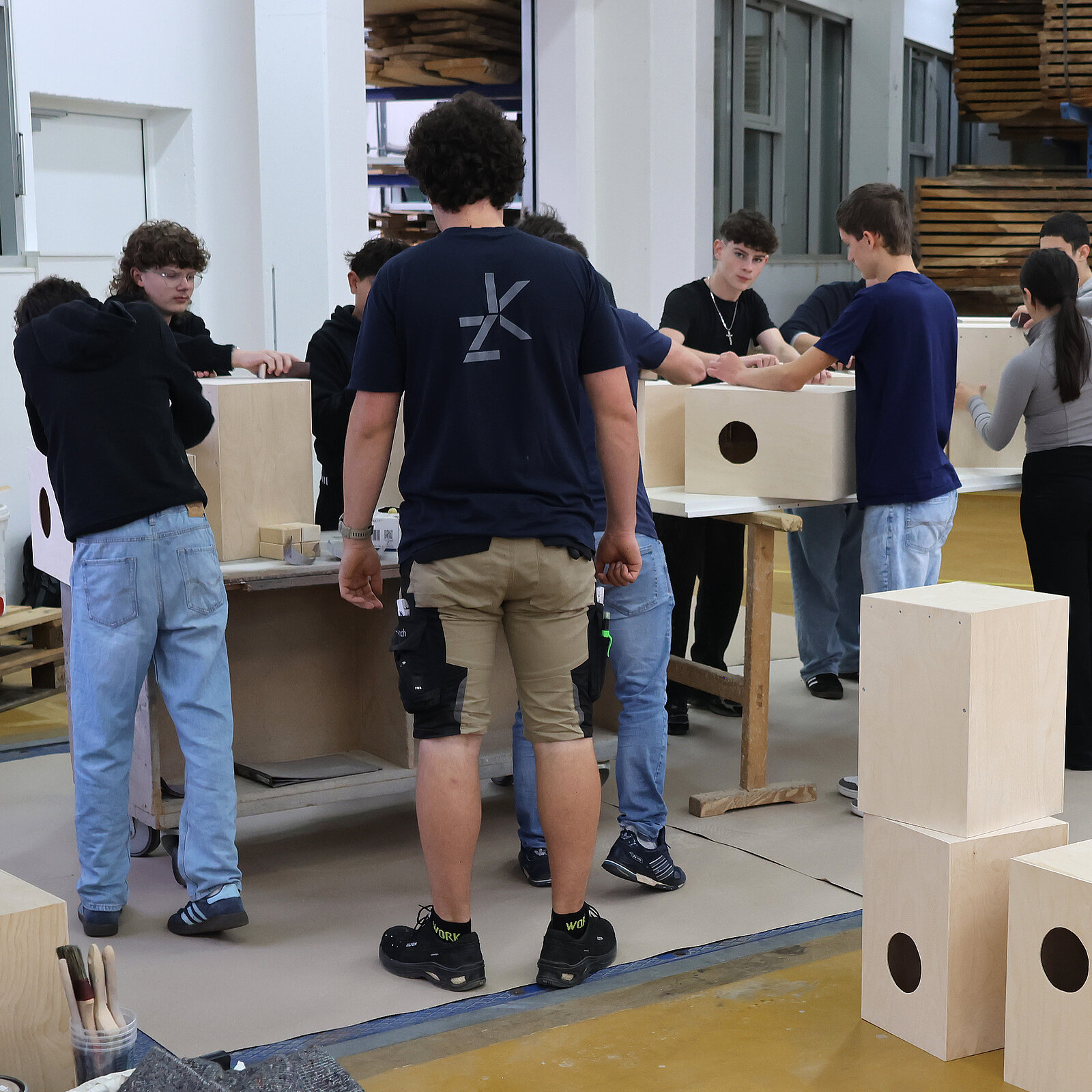 Gruppe von Jugendlichen arbeitet in einer Werkstatt an Holzprojekten. Sie stehen um Tische und bearbeiten Kisten aus Holz. Die Atmosphäre ist konzentriert, und die jungen Menschen scheinen in ihre Aufgaben vertieft.
