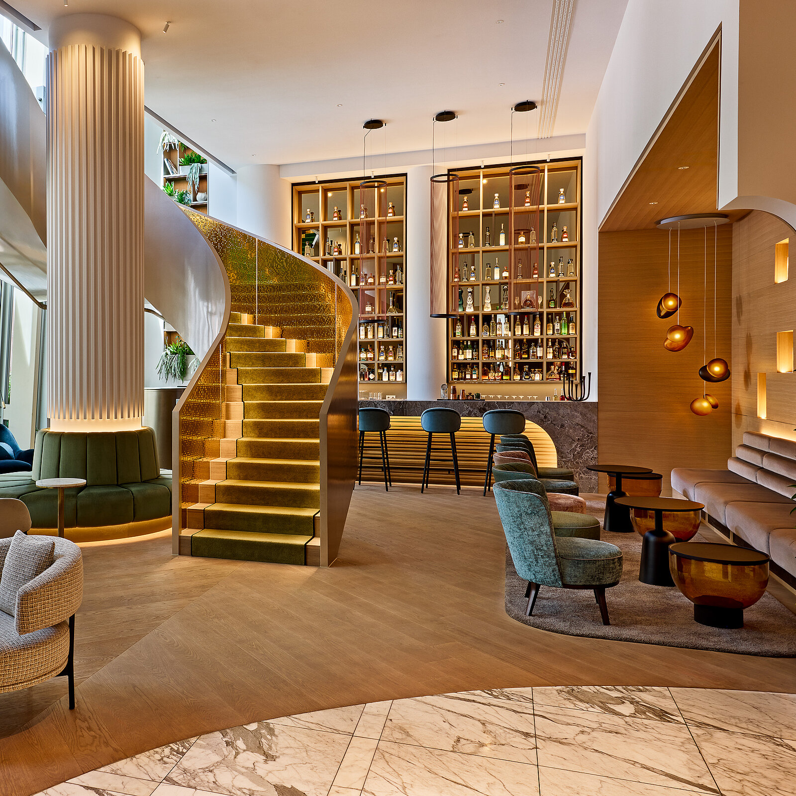 Ein eleganter Lounge-Bereich mit einer geschwungenen Treppe, die zu einer Galerie führt. Die Treppe ist beleuchtet und von bequemen Sitzgelegenheiten umgeben. Im Hintergrund befindet sich eine stilvolle Bar mit Regalen voller Flaschen und blauer Barhocker.