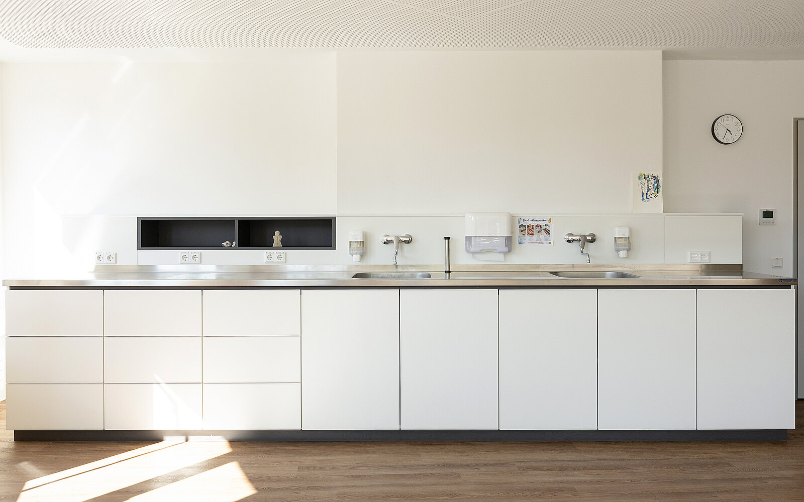 Eine moderne, minimalistische Küche mit weißen Schränken und Arbeitsplatte. Drei Wasserhähne sind in der Mitte installiert. Wände und Boden sind schlicht gestaltet.