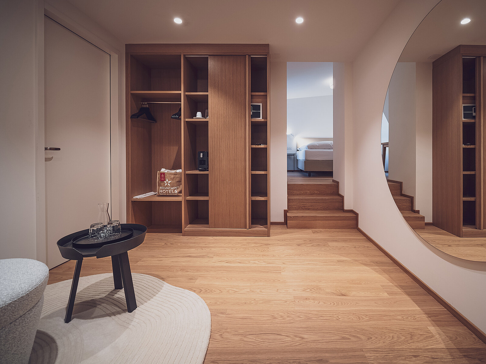 Moderner Ankleideraum mit Holzregalen, einige Fächer sind leer, während in anderen Kleidungsstücke und Kaffeekapseln zu sehen sind. Ein runder Tisch mit einer Karaffe steht auf einem Teppich. Eine Treppe führt zu einem Schlafzimmer im Hintergrund. Ein großer runder Spiegel an der Wand.