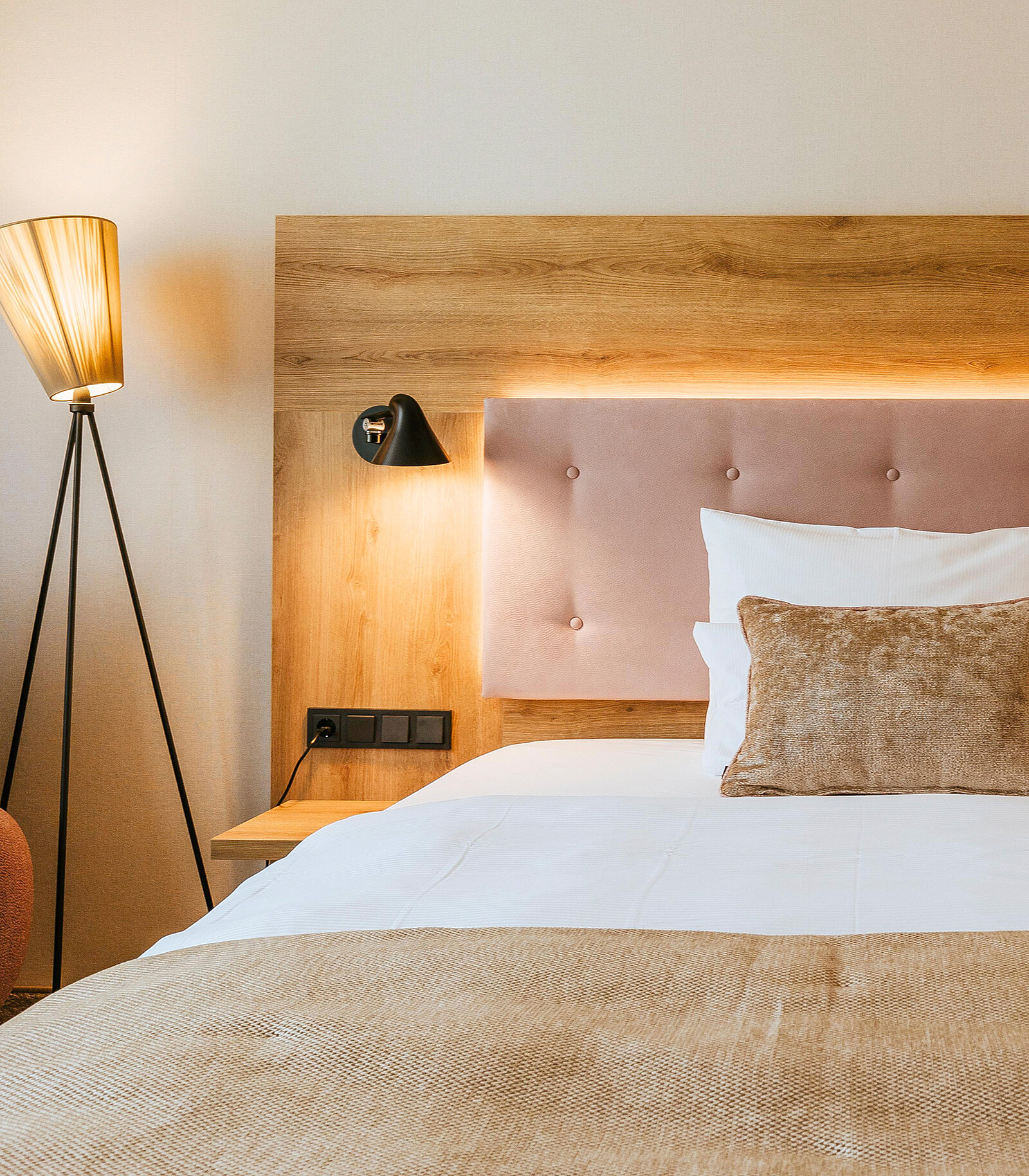 Das Bild zeigt ein modernes Schlafzimmer mit einem bequemen Bett, das eine gepolsterte Kopfteilwand hat. Neben dem Bett steht ein Nachttisch mit einer Lampe. Eine dekorative Stehlampe steht in der Ecke des Raumes, der in warmen Farben gehalten ist.