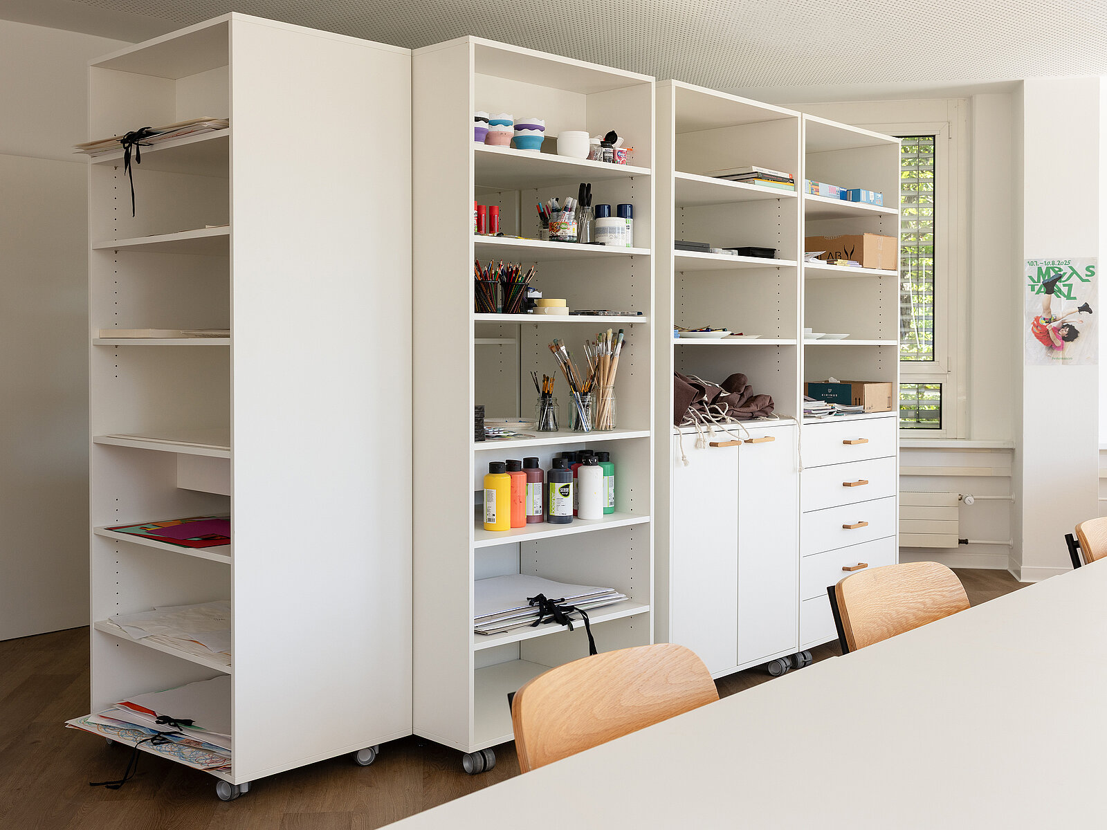 Kunstmaterialien auf weißen Regalen in einem gut beleuchteten Raum. Es sind Farben, Pinsel, Stifte und andere Bastelutensilien organisiert. Vor den Regalen steht ein Tisch mit Holzstühlen. Ein Fenster an der rechten Seite lässt Tageslicht herein.
