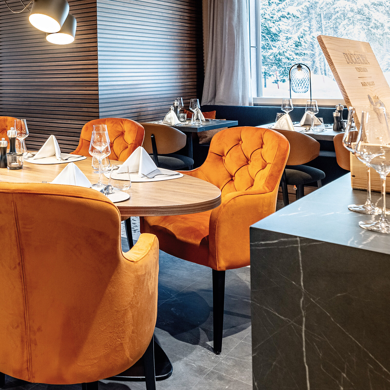 Zieflekoch hotel piz badus Andermatt Schweiz Restaurant Frühstücksraum nussbaum wandbild berge steinwand kamin nussbaum lamellen polstersessel gedeckter Tisch Hängeleuchten winter