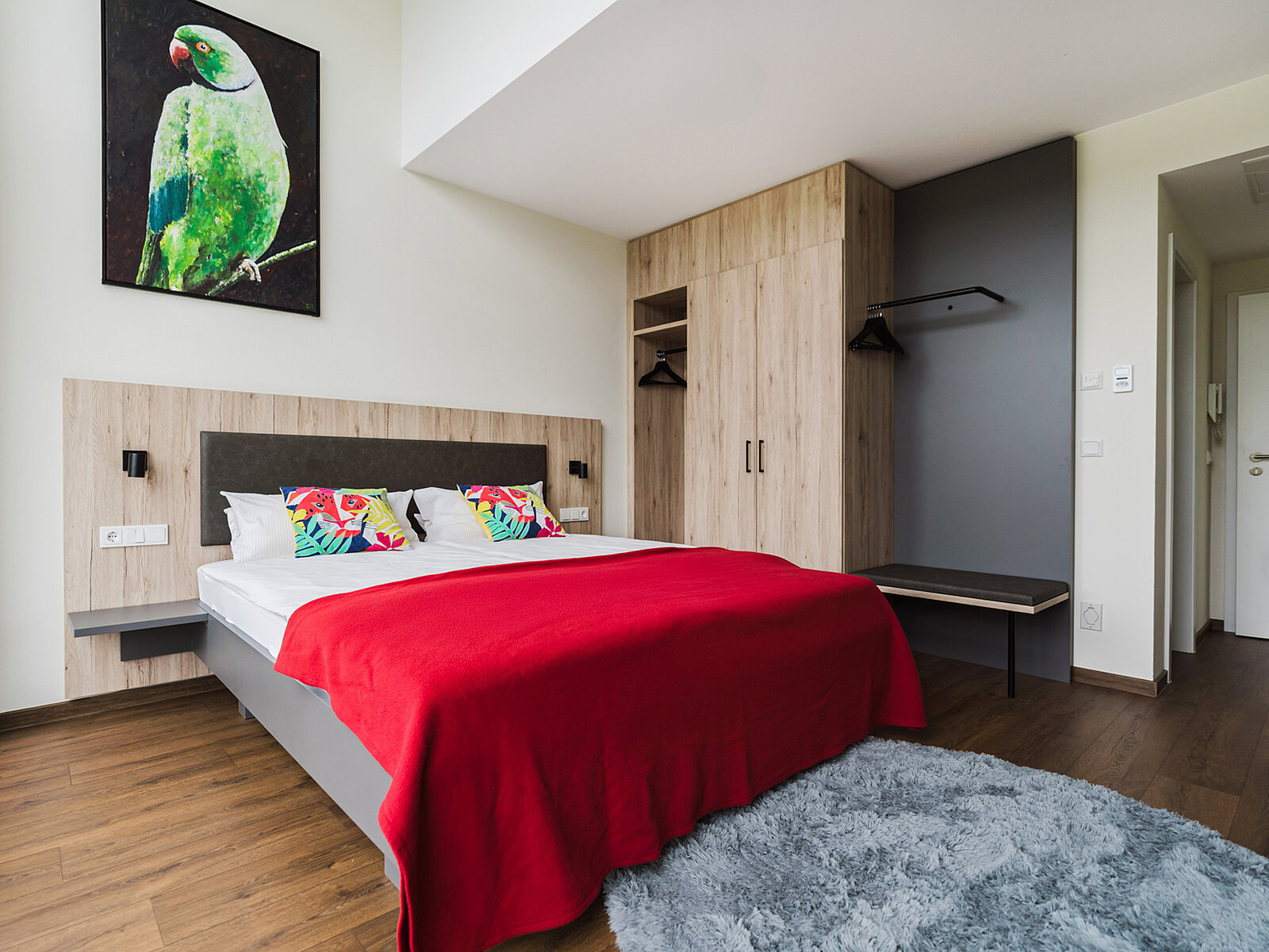 Ein modernes Schlafzimmer mit einem Doppelbett, roter Decke und bunten Kissen. An der Wand hängt ein Gemälde eines Papageis. Holzfußboden mit einem grauen Teppich. Wandschrank und Haken an der Seitenwand.