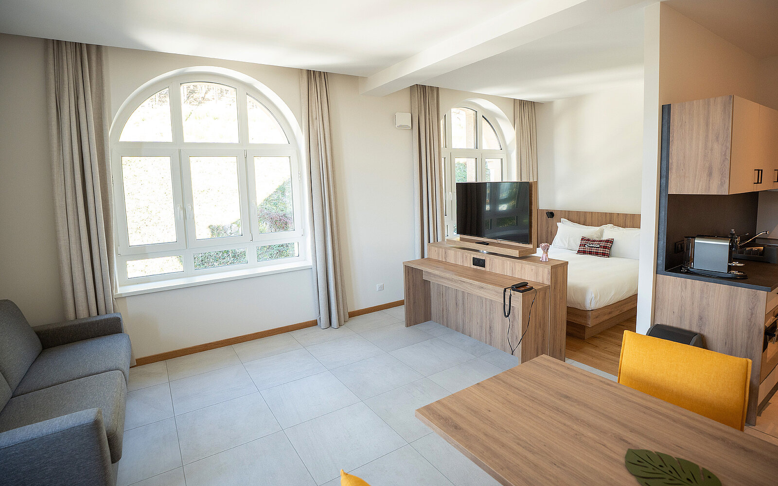 Helles Apartment mit großen Fensterbögen, modernem Wohnzimmer, grauem Sofa, Holztisch und gelben Stühlen. Offene Küche rechts, gegenüber ein Bett mit Fernseher. Neutrale Farbtöne und minimalistisches Design.