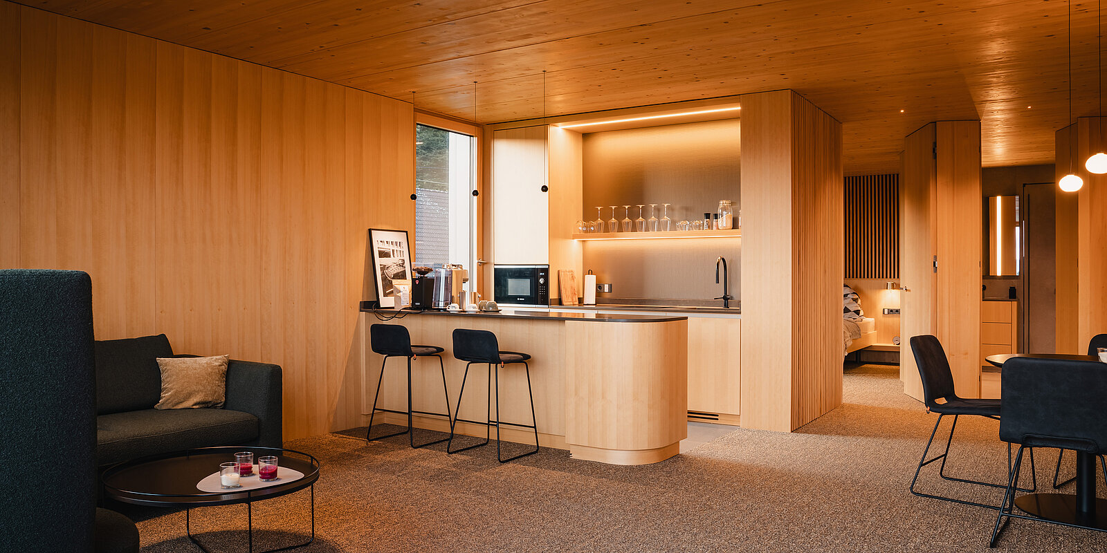 Moderne Küche in Holzoptik mit einer Mittelinsel und Barhockern. Links ein Sofa mit einem kleinen Tisch. Rechts ein Esstisch mit Stühlen. Warme, einladende Beleuchtung und offene Gestaltung betonen das elegante, minimalistische Design des Raumes.