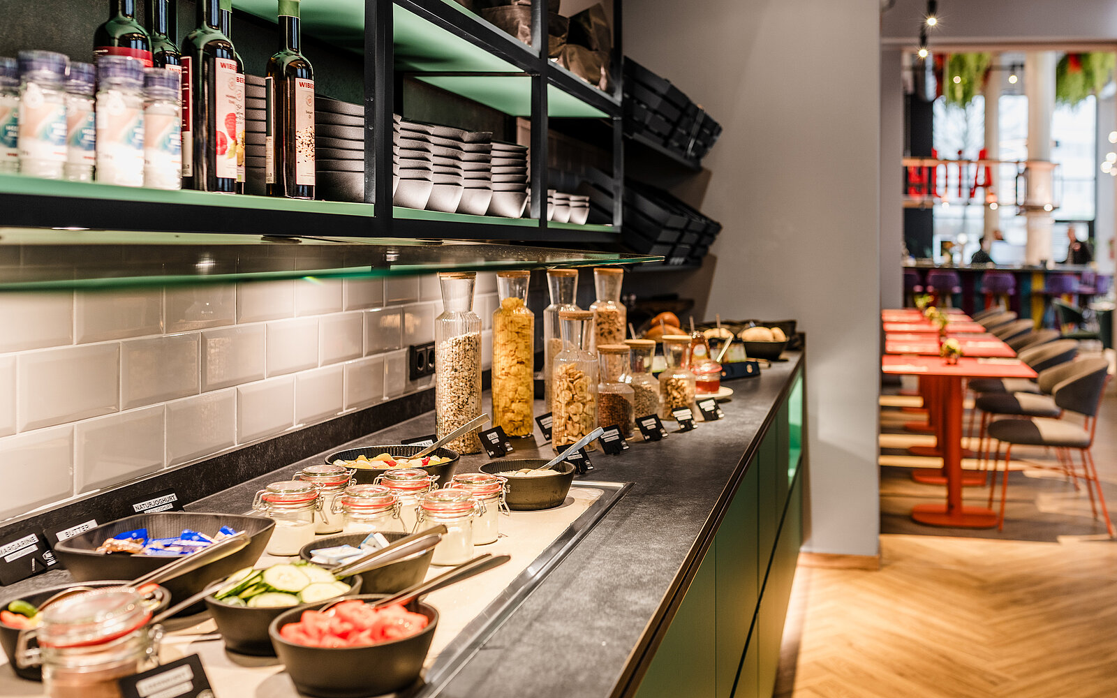 Ein Frühstücksbuffet mit einer Auswahl an Müsli, Joghurt und frischen Zutaten in Schalen. Im Hintergrund sind Tische und Stühle in einem Essbereich erkennbar.