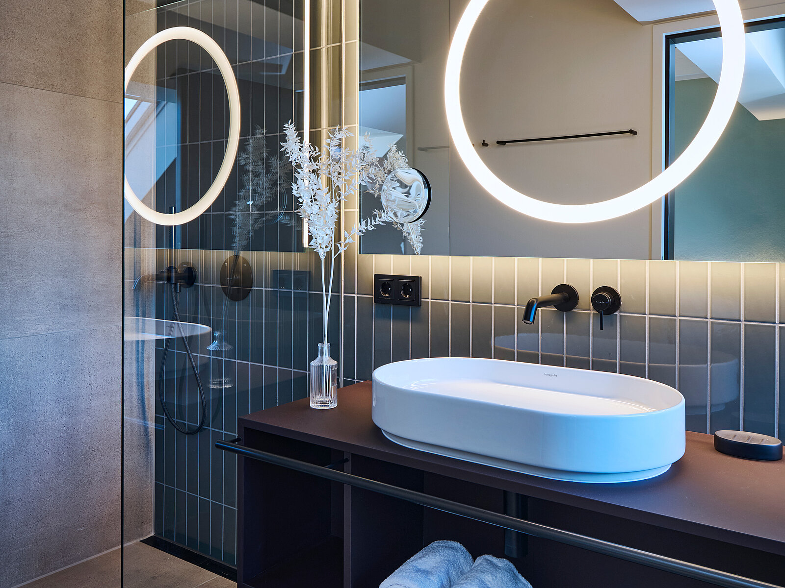 Ein modernes Badezimmer mit einem ovalen Waschbecken auf einer dunklen Ablage. Darüber befindet sich ein großer, beleuchteter, runder Spiegel. Die Wand ist mit schmalen Fliesen verkleidet. Ein kleines Glasgefäß mit dekorativen Zweigen steht neben dem Waschbecken.