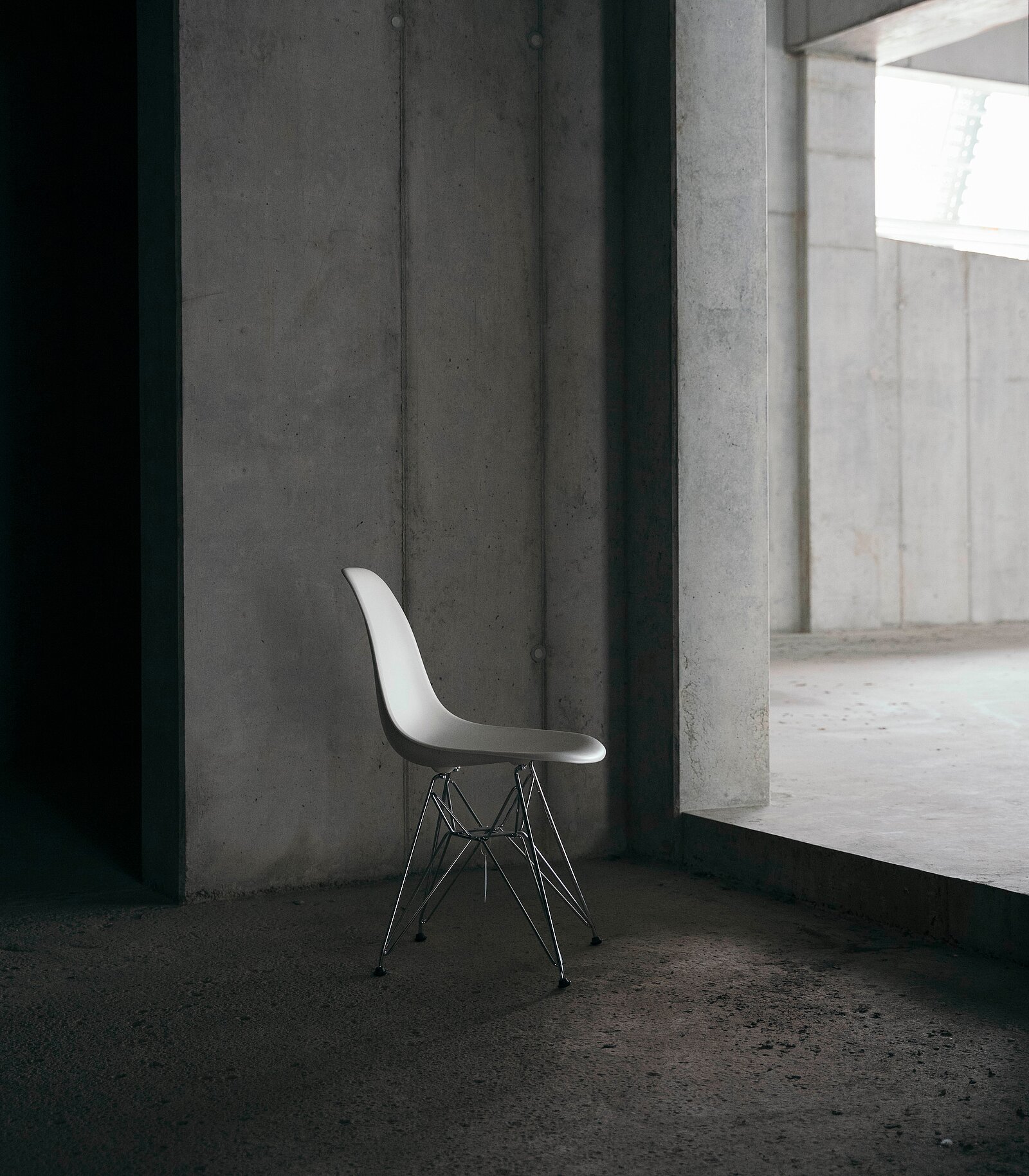 Ein einzelner, moderner, weißer Stuhl mit einem metallischen Gestell steht in einem minimalistischen Raum mit grauen Wänden und Betonboden. Das Licht fällt sanft durch ein Fenster und hebt die Schlichtheit des Möbelstücks hervor.