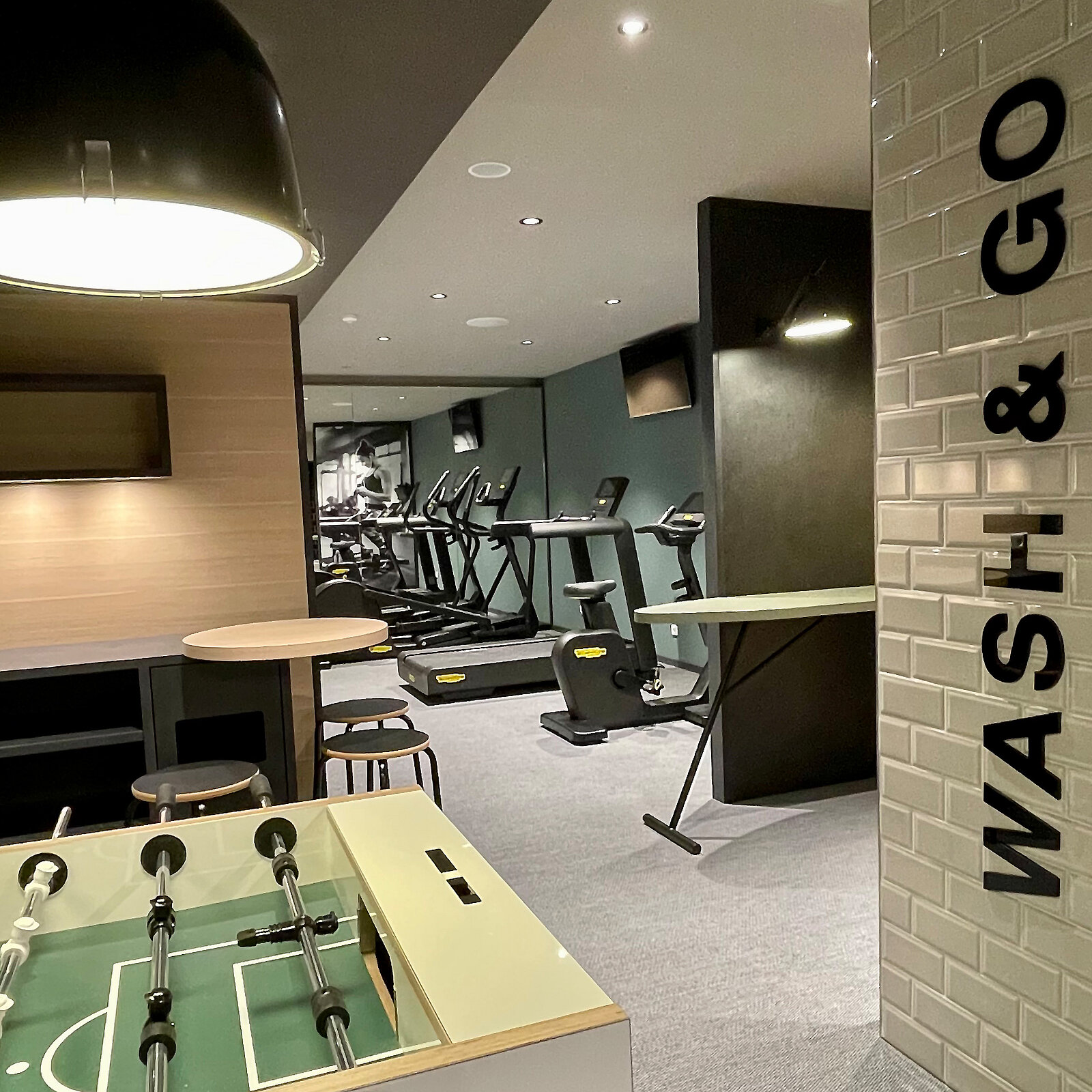 Ein moderner Fitnessraum mit Heimtrainern und einem Kicker-Tisch im Vordergrund. An der Wand steht der Schriftzug "WASH & GO". Die Einrichtung ist schlicht und einladend, mit einer hellen Beleuchtung und neutralen Farben.