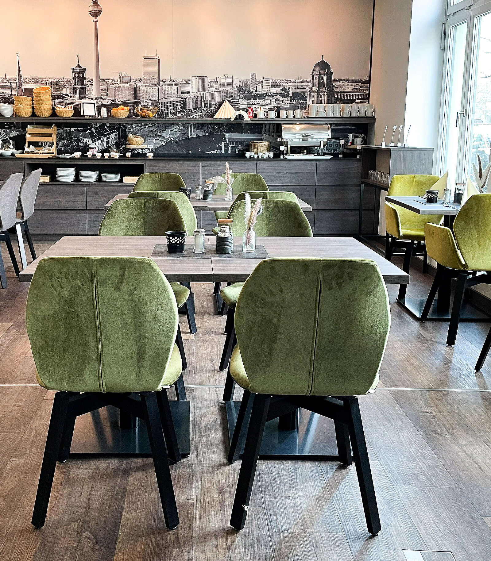 Moderner Frühstücksraum mit mehreren Tischen und grün gepolsterten Stühlen. Im Hintergrund ein Buffet mit Brotkörben und Getränken vor einem großformatigen Schwarz-Weiß-Bild einer Stadtansicht.