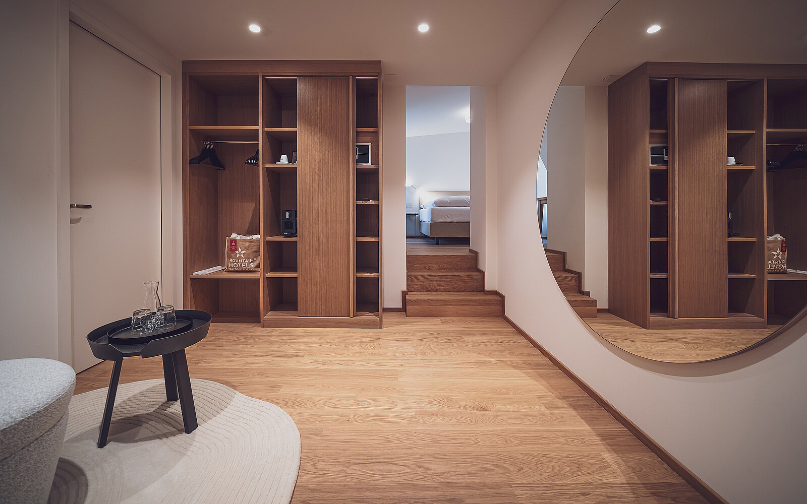 Moderner begehbarer Kleiderschrank mit Holzregalen, Spiegel und rundem Tisch. Im Hintergrund führt eine kleine Treppe zu einem Schlafzimmer. Dezente Beleuchtung sorgt für eine einladende Atmosphäre.
