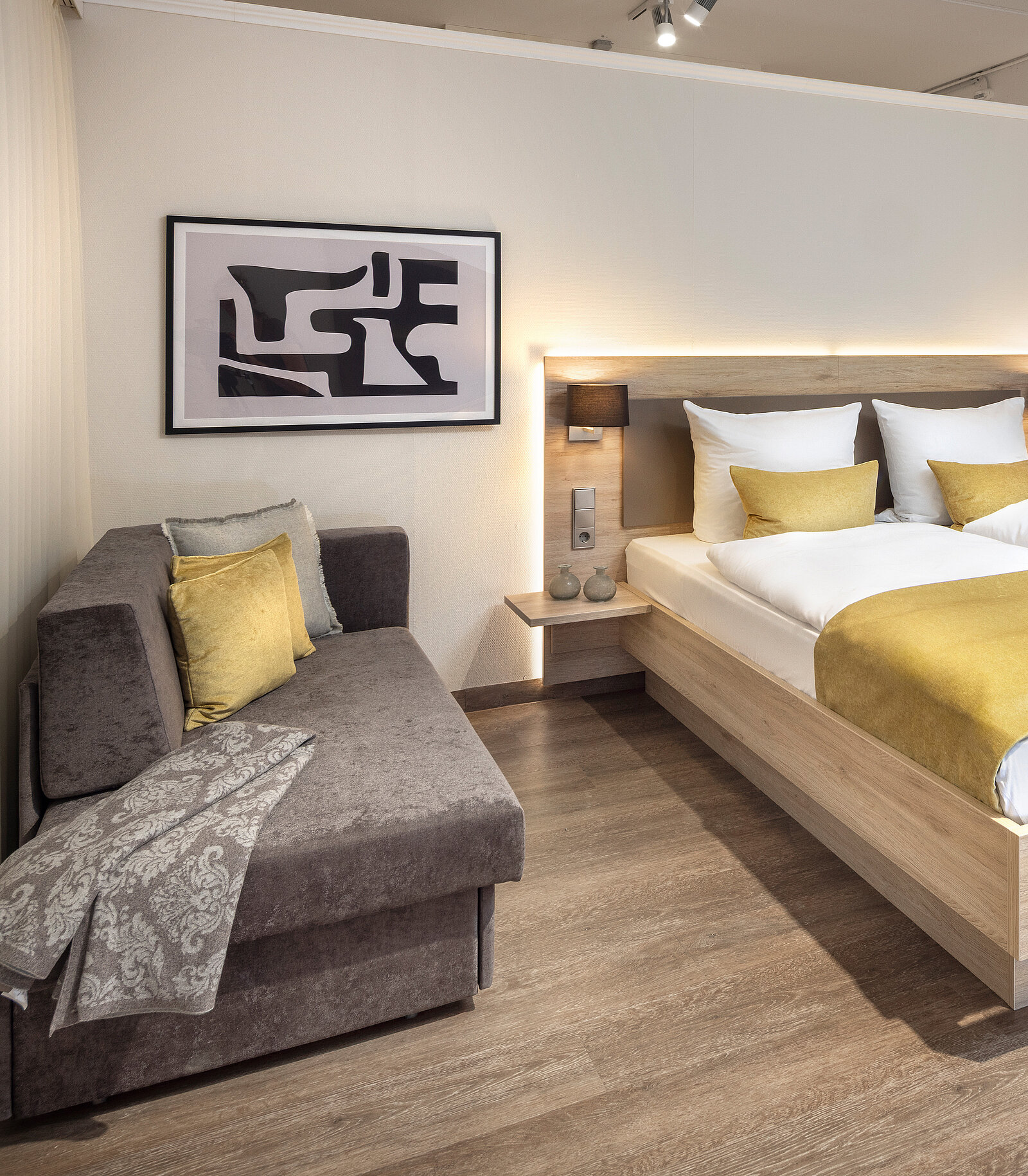 In einem modernen Schlafzimmer befinden sich ein Bett mit hellen Kissen und einer goldenen Decke sowie eine graue Couch mit einem gelben Kissen. An der Wand hängt ein abstraktes Kunstwerk. Der Boden ist aus Holz, und die Beleuchtung ist warm und einladend.
