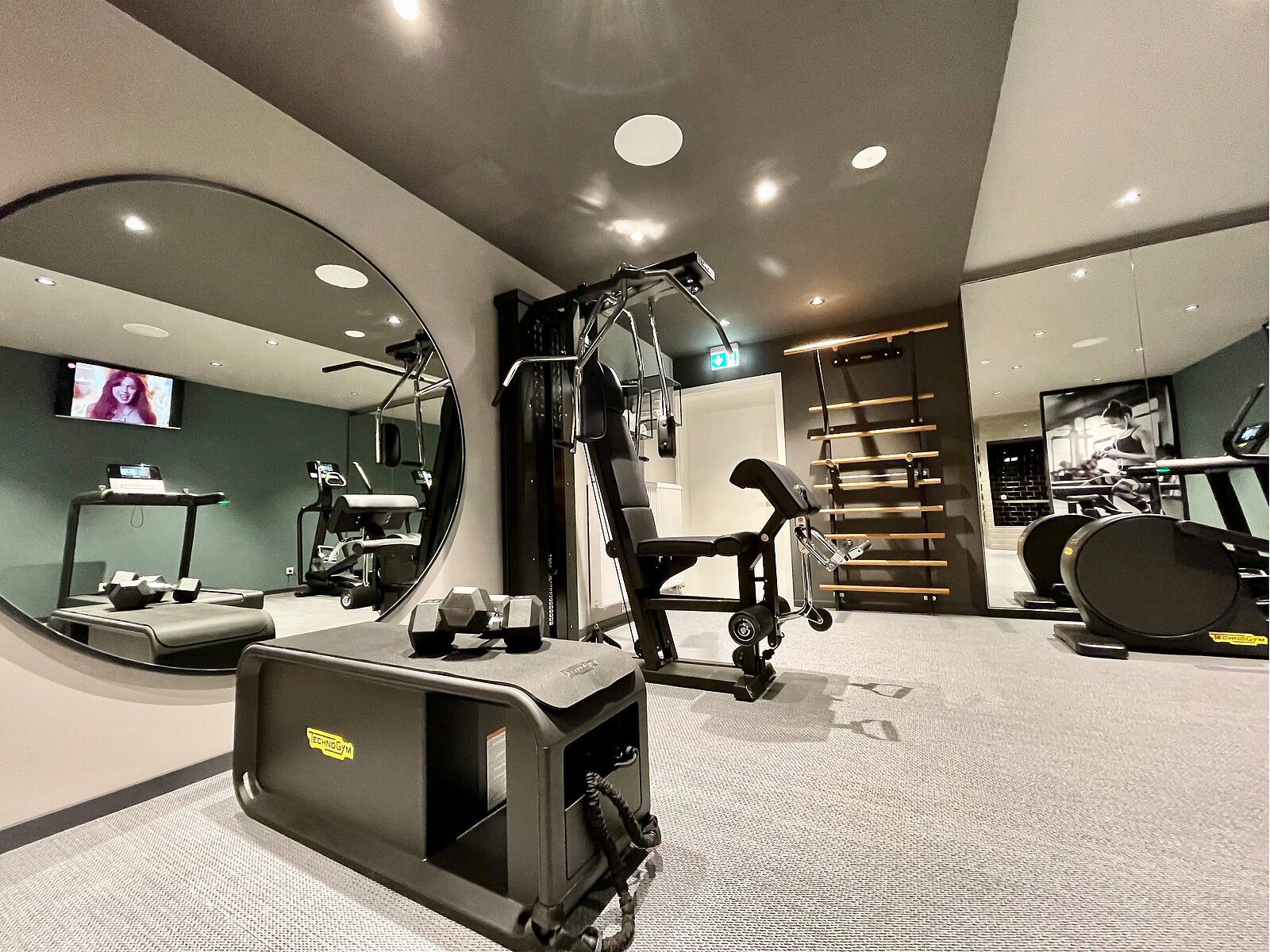 Ein moderner Fitnessraum mit Kraftgeräten. Auf der linken Seite befindet sich ein Laufband, daneben ein Spiegel. An der Wand hängt ein Regal für Trainingsgeräte. Im Hintergrund ist ein Fernseher zu sehen, der eine Person zeigt. Der Boden ist mit grauem Teppich ausgelegt.