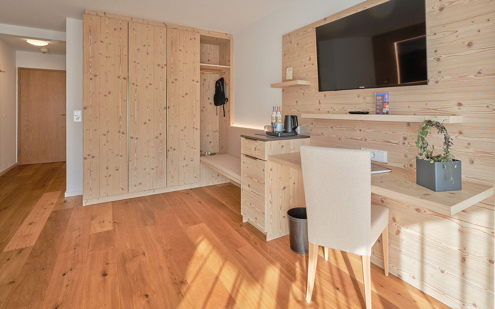 Helles Hotelzimmer mit Holzmöbeln, inklusive großem Kleiderschrank, Schreibtisch mit Stuhl und Wandfernseher. Teppichloser Holzboden. Zimmer wirkt modern und aufgeräumt.