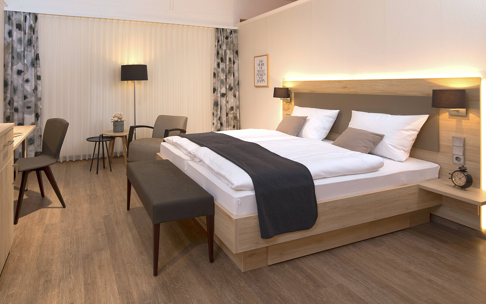 Das Bild zeigt ein modernes Hotelzimmer mit einem Doppelbett, das mit weißen Bettlaken und einem grauen Überwurf dekoriert ist. Neben dem Bett steht eine Holzbank. Im Hintergrund gibt es einen Tisch mit zwei Stühlen und Vorhänge, die Tageslicht filtern.