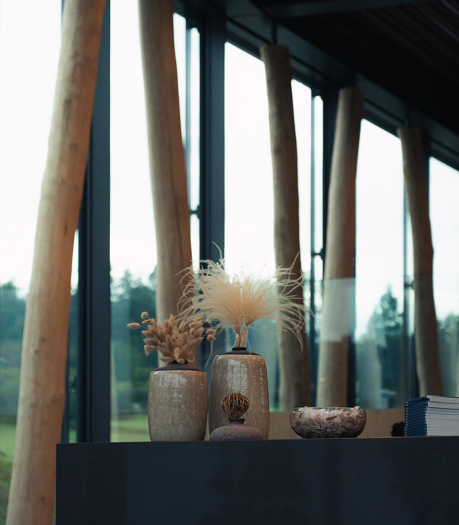 Eine minimalistische Inneneinrichtung zeigt drei dekorative Vasen auf einer dunklen Oberfläche. Zwei Vasen enthalten getrocknete Pflanzen, während eine kleinere Schale daneben steht. Im Hintergrund sind große Fenster mit Blick auf Bäume und einfallendes Tageslicht zu sehen.