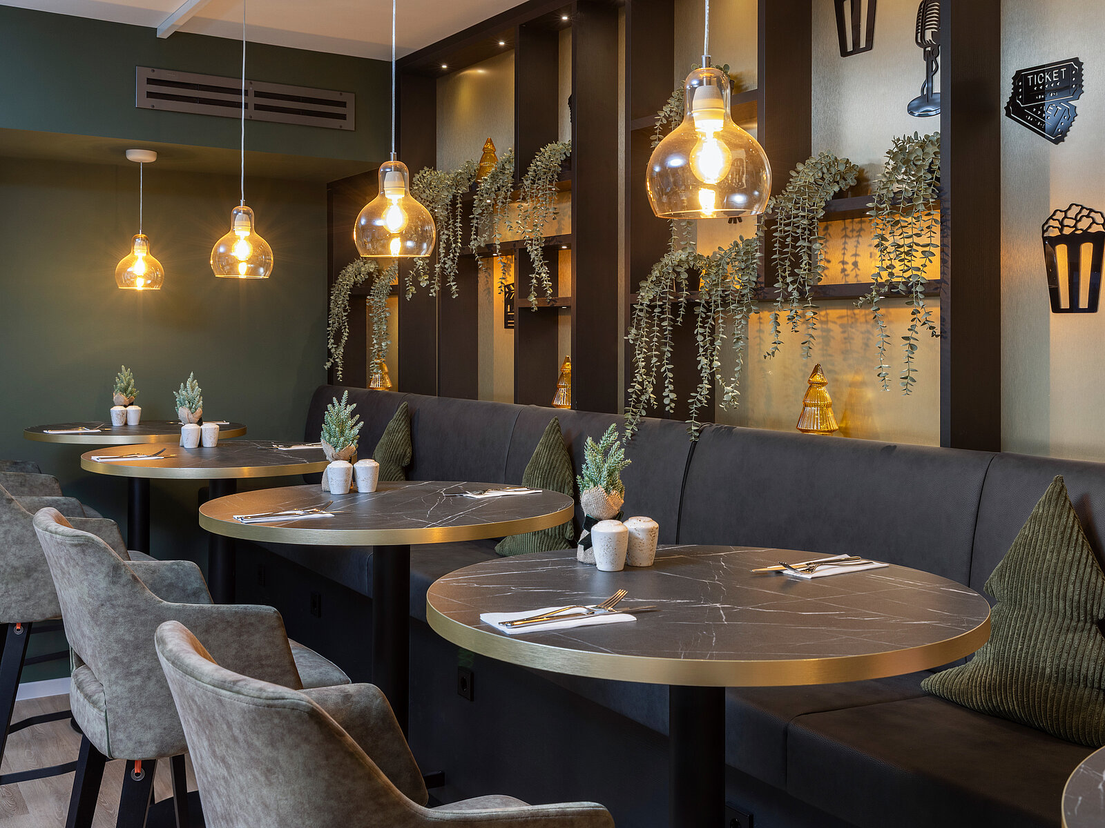 A cozy restaurant interior featuring round marble tables surrounded by upholstered chairs. Soft lighting from pendant lamps illuminates the space, and small potted plants add a touch of greenery. The walls are decorated with shelves holding decorative items.