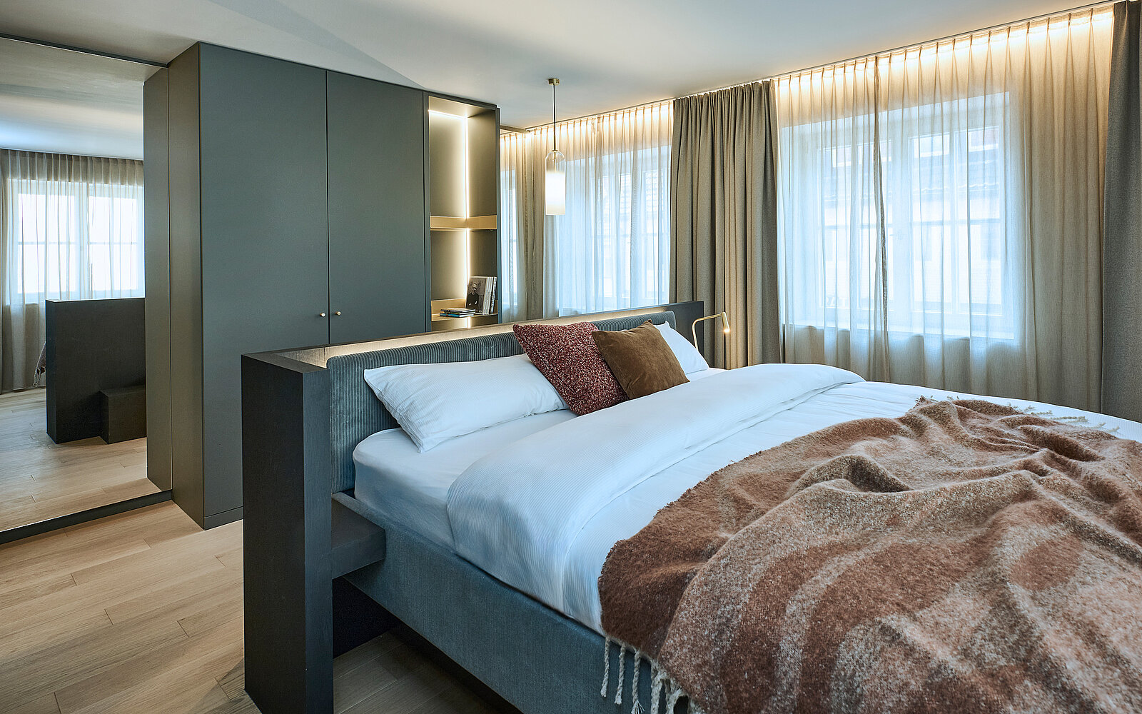 Modernes Schlafzimmer mit einem großen Bett, weißen Kissen und einer braunen Decke. Der Raum ist hell erleuchtet, hat einen großen Kleiderschrank und lange Vorhänge vor den Fenstern. Eine dezente Hängelampe sorgt für zusätzliche Beleuchtung.