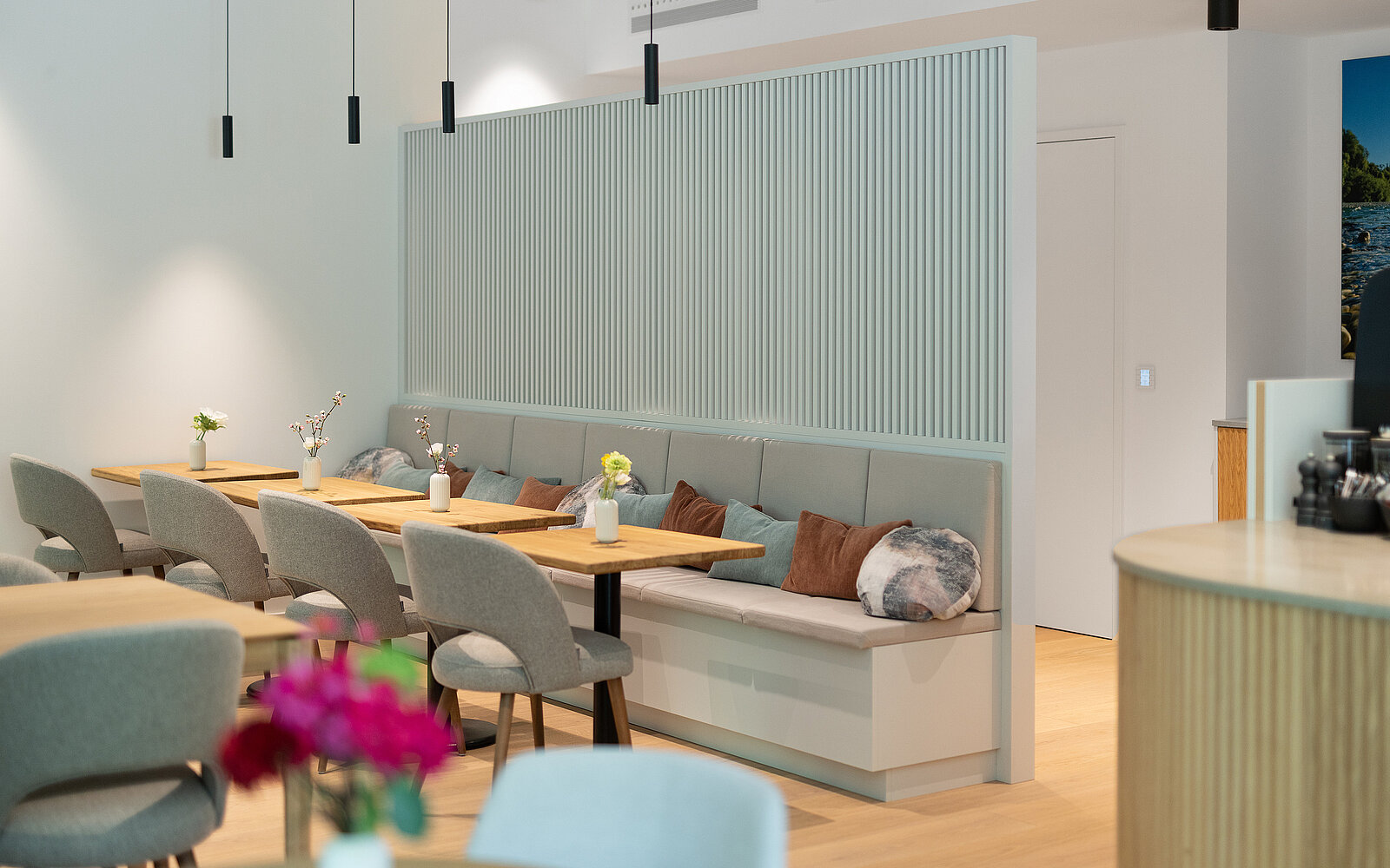 Moderner Essbereich in einem Café oder Restaurant mit hellen Holztischen und grauen Polsterbänken. Der Raum ist mit minimalistischen Vasen und Blumen dekoriert. Eine hölzerne Trennwand teilt den Bereich. Hängeleuchten sorgen für stimmungsvolles Licht.