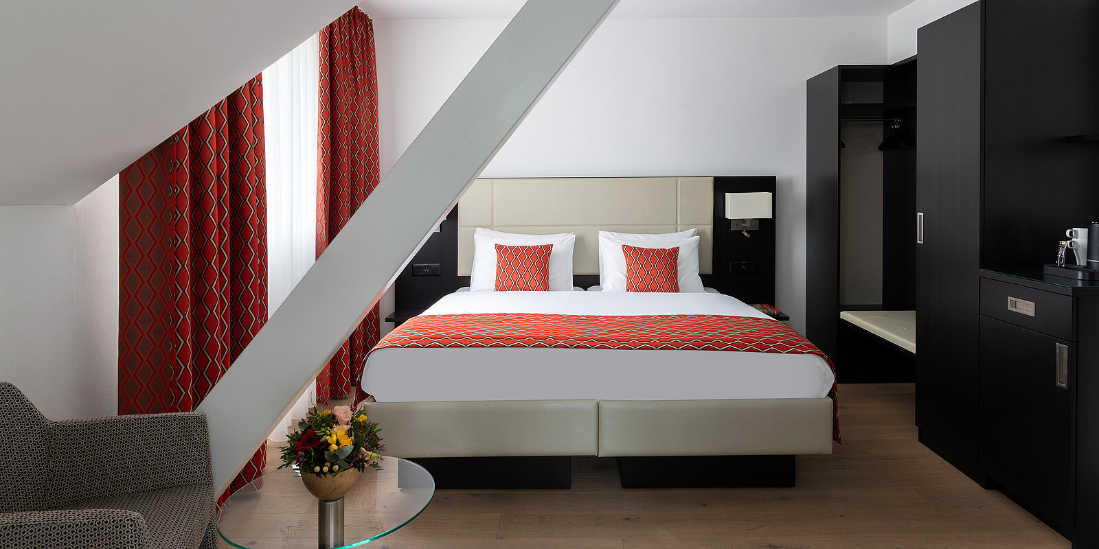 Modernes Hotelzimmer mit großem Doppelbett, weißen und roten Akzenten. Eine gemusterte Decke und Kissen in Rot, Vorhänge passend. Ein elegantes Sofa und ein Glastisch mit Blumen daneben. Schrank und Minibar im Hintergrund. Helle Wände und Holzboden.