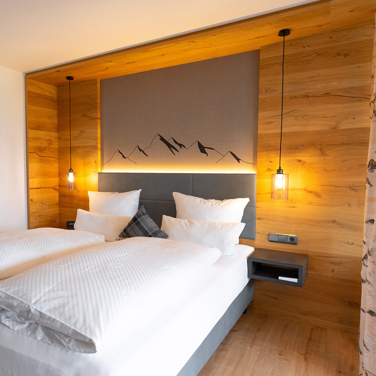 Ein modernes Schlafzimmer mit einem großen Doppelbett, hellen Kissen und einer stylischen Kopfteilwand. Die Wände sind aus Holz, und es gibt eine sanfte Beleuchtung, die eine gemütliche Atmosphäre schafft. Im Hintergrund ist ein minimalistisches Bergmuster zu sehen.