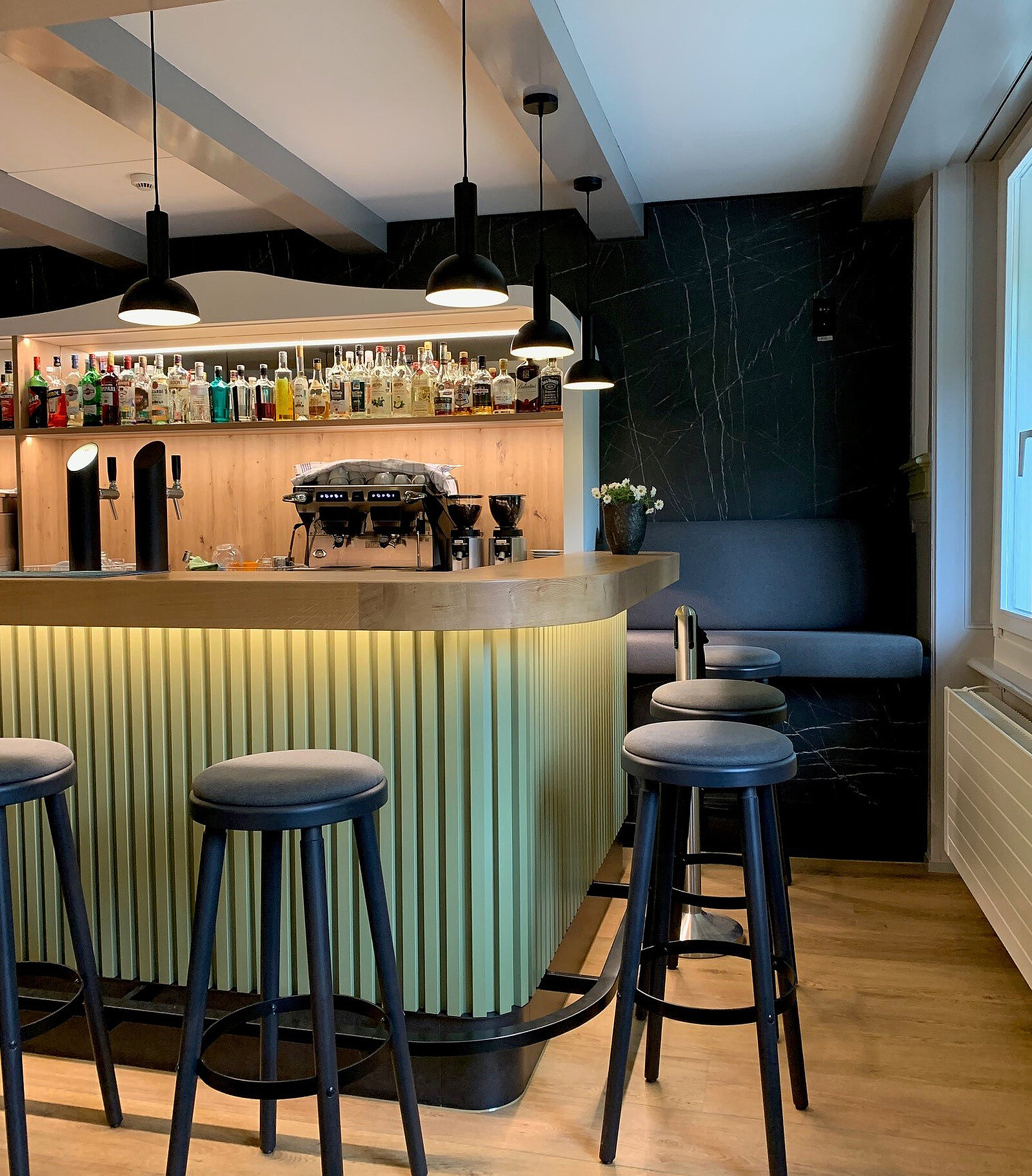 Das Bild zeigt eine stilvolle Bar mit einem gegrünten Bartresen und modernen, runden Barhockern. Im Hintergrund stehen Regale mit einer Vielzahl von Spirituosen, und die sanfte Beleuchtung schafft eine einladende Atmosphäre. Ein Sitzbereich an der Wand ergänzt das Ambiente.