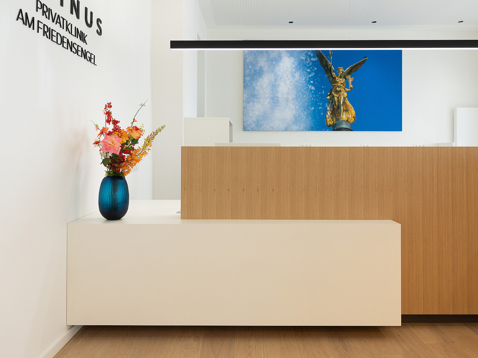 Eine moderne Rezeption mit einem Holztresen. Auf der linken Seite steht eine Vase mit bunten Blumen auf einem weißen Tisch. An der Wand hängt ein Bild von einer goldenen Engelsstatue vor blauem Himmel.