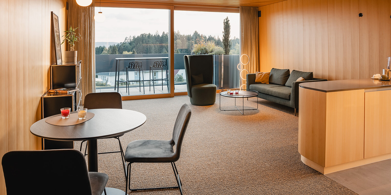 Modernes Wohnzimmer mit großen Fensterfronten und Ausblick auf einen Balkon. Links steht ein runder Tisch mit zwei Stühlen, rechts eine schwarze Couch und ein runder Couchtisch. Die Wände sind mit Holz verkleidet, was eine warme Atmosphäre schafft.