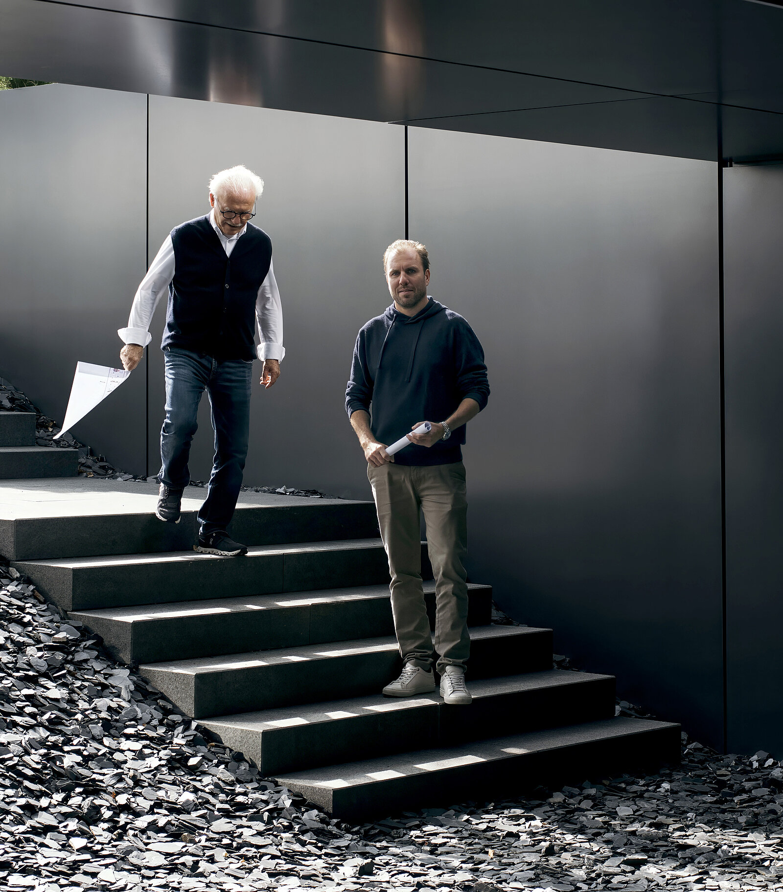Zwei Männer stehen auf einer Treppe, die aus dunklen Steinen besteht. Der ältere Mann mit grauen Haaren trägt eine Weste und hält ein Blatt Papier, während der jüngere Mann in einem dunklen Pullover daneben steht und einen Stift in der Hand hält.