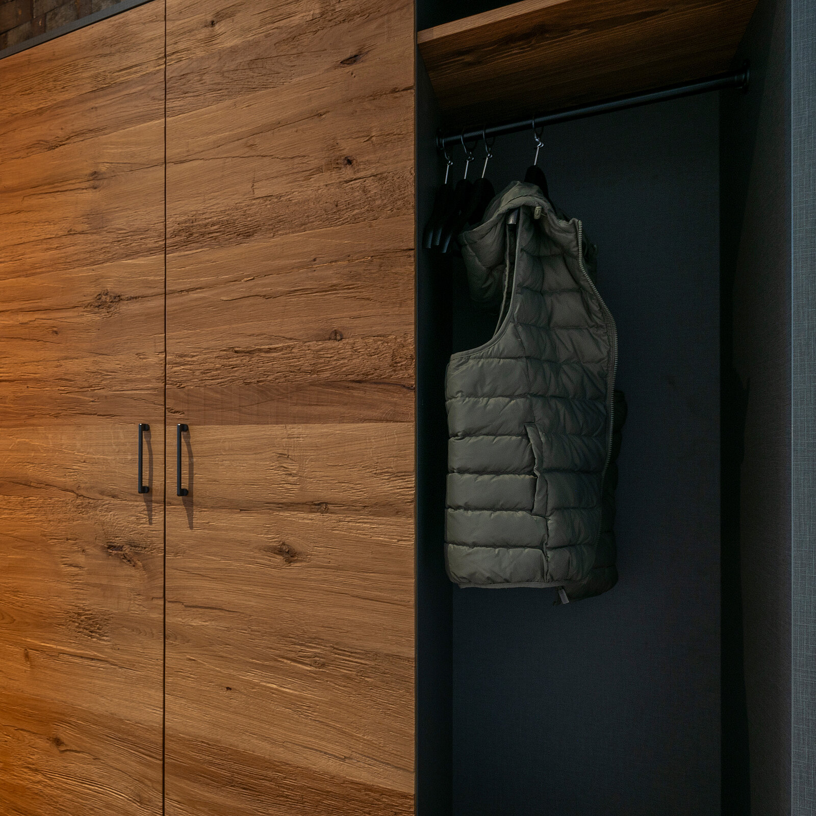 Ein moderner Kleiderschrank aus Holz mit zwei Türflügeln. Links ist eine graue Weste auf einem Kleiderbügel aufgehängt. Die Holzoberfläche wirkt glatt und elegant, die Umgebung ist schlicht und neutral gestaltet.