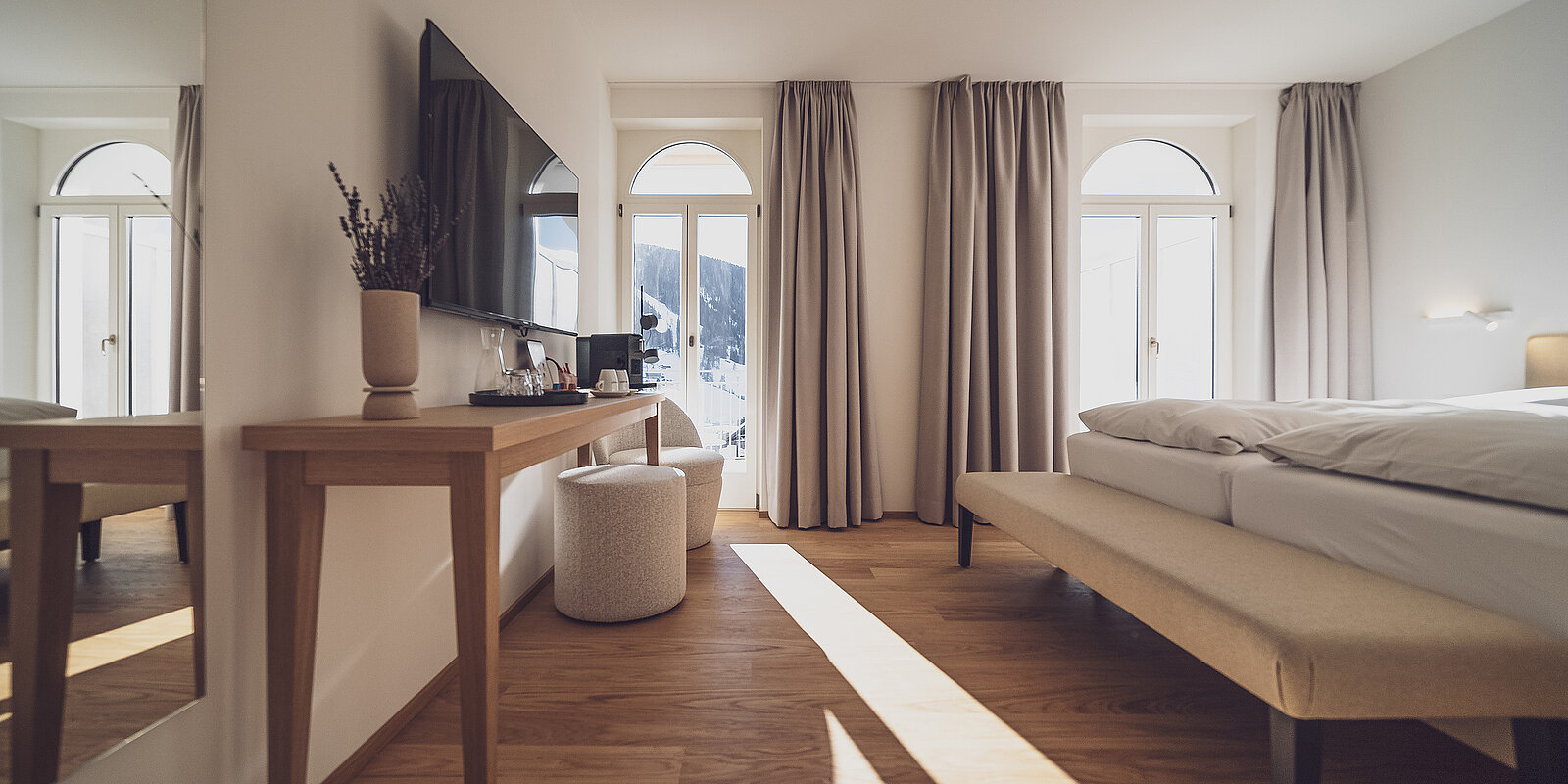 Helles Hotelzimmer mit großem Bett, Holzfußboden, Flachbildfernseher und langem Schreibtisch. Vorhänge an Fenstern, durch die Sonnenlicht einfällt. Minimalistisch eingerichtet.
