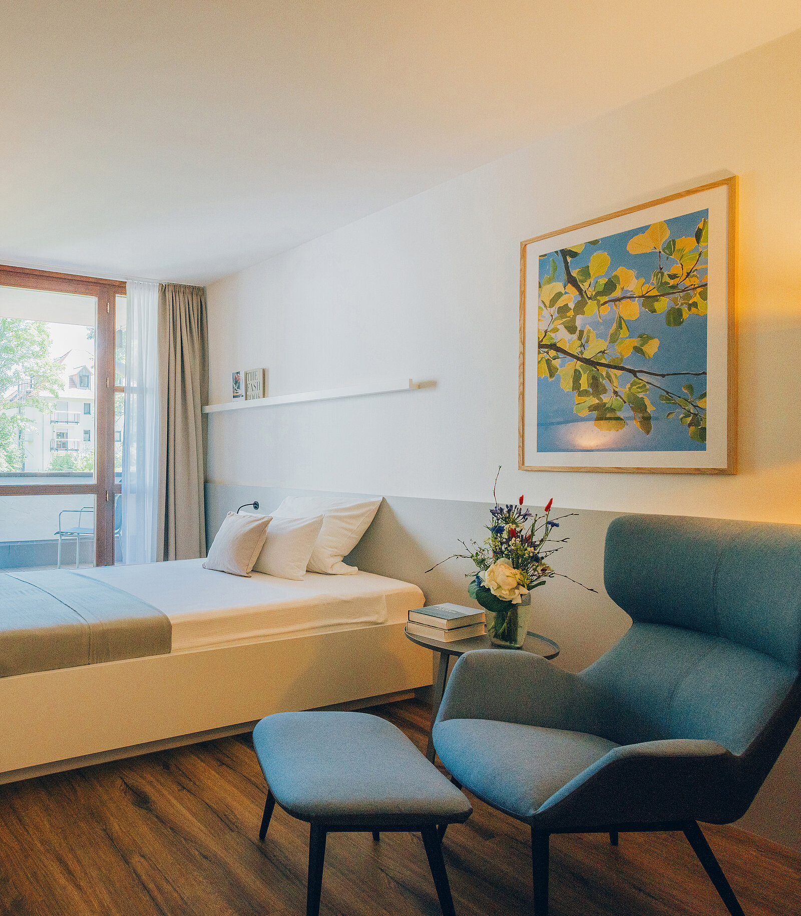 Helles, modernes Hotelzimmer mit Holzfußboden. Links steht ein Doppelbett, daneben ein kleiner Tisch mit Blumen. Rechts ein grauer Sessel mit Hocker. An der Wand hängt ein Bild mit Naturmotiv. Fenster mit Blick auf grüne Bäume.