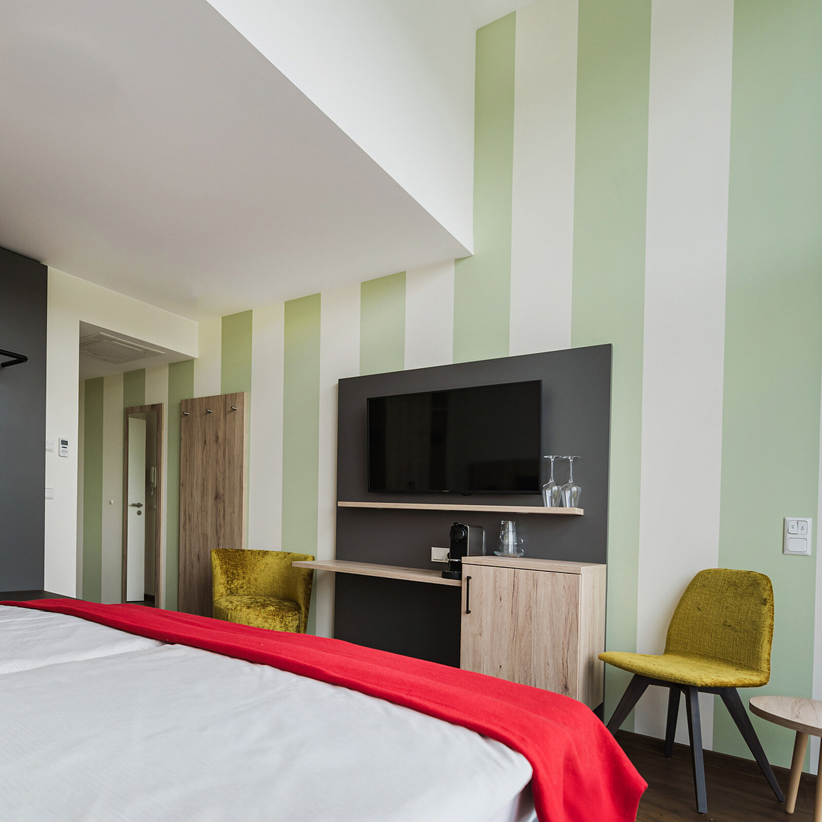 Ein modernes Hotelzimmer mit gestreiften Wänden, einem Doppelbett mit rotem Überwurf, einem Flachbildfernseher an der Wand, einem kleinen Tisch mit Wasserkocher und Tassen, sowie zwei gelben Stühlen.