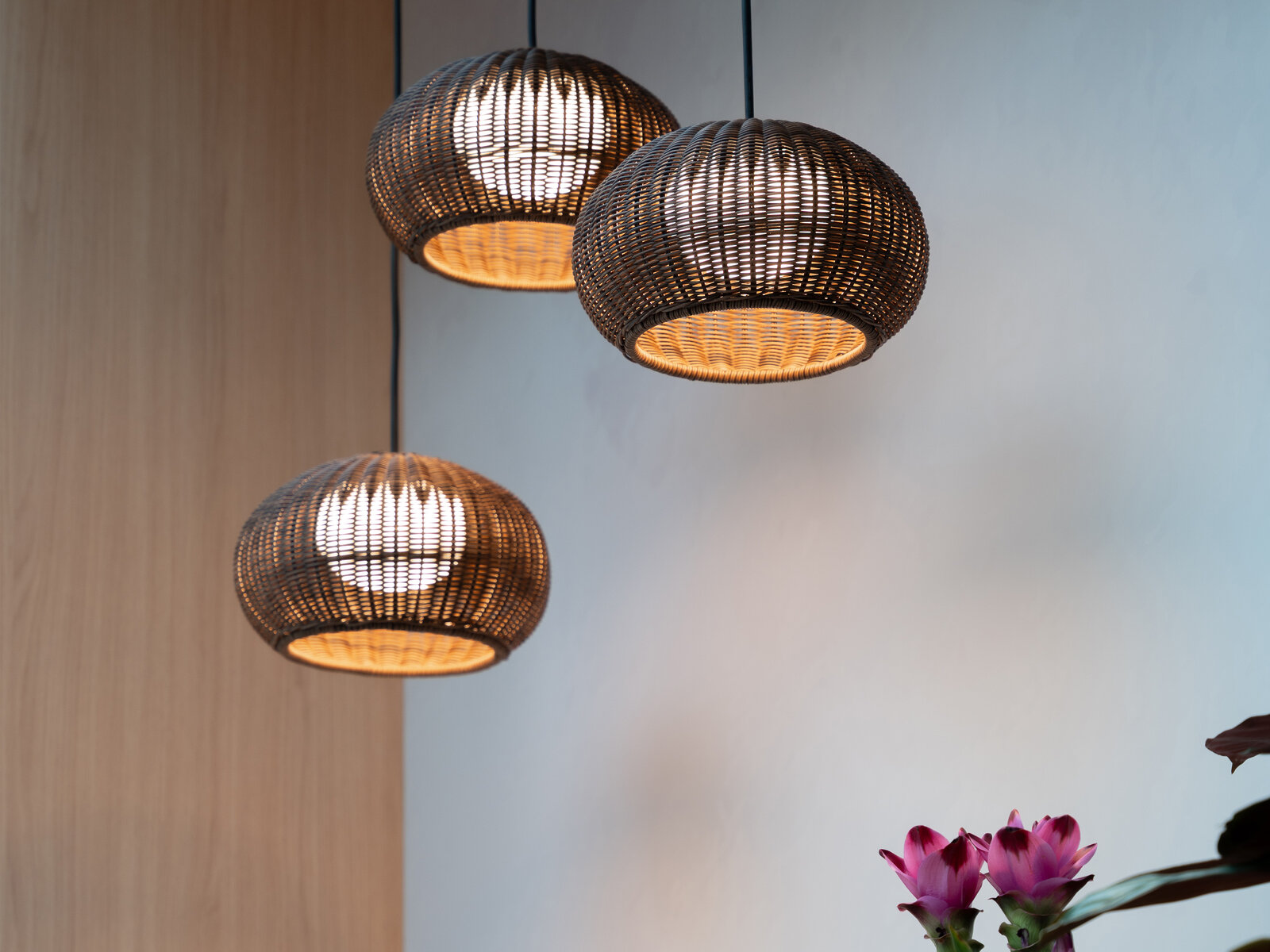 Three woven rattan pendant lights hang against a light wall, with pink flowers visible in the bottom right corner.