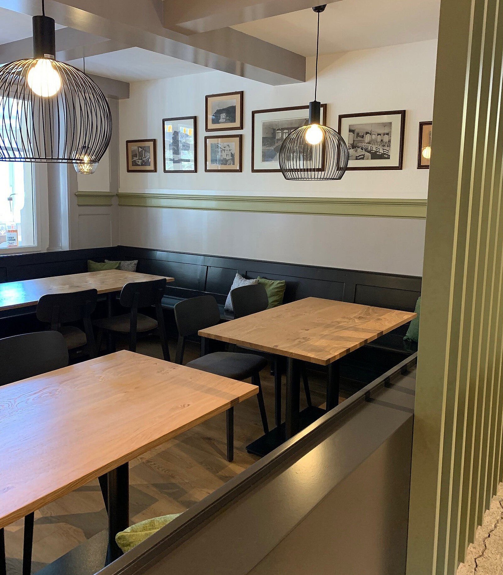 Ein gemütlicher Restaurantbereich mit Holztafeln und schwarzen Stühlen. An der Wand hängen Bilder in Rahmen. Es gibt Pendelleuchten, die den Raum sanft beleuchten, und eine grüne Wandverkleidung. Die Atmosphäre wirkt einladend und freundlich.