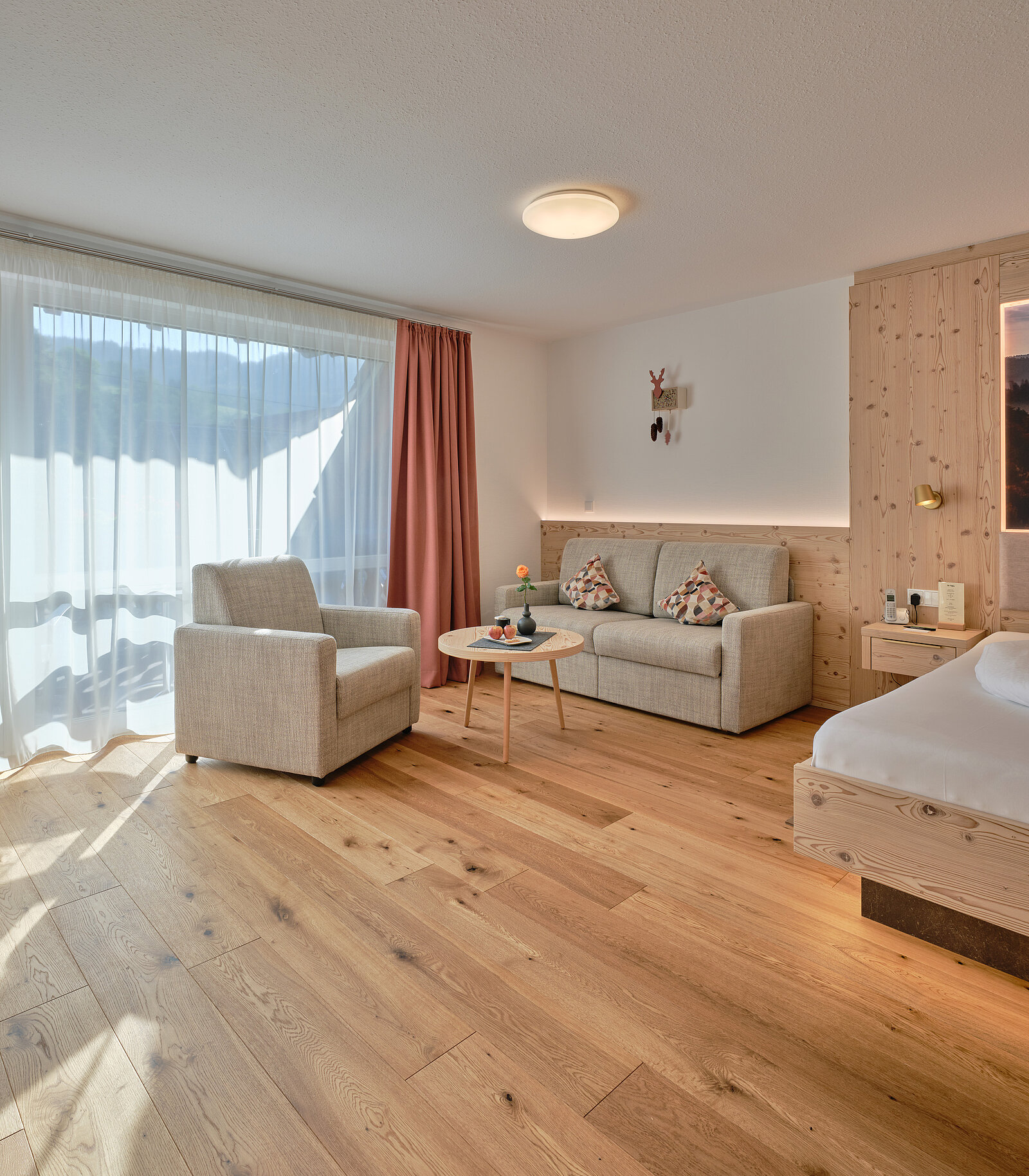 Helle Hotelzimmer mit Holzboden, Doppelbett, Sofa und Sessel. Ein kleiner runder Tisch steht vor dem Sofa. Tageslicht strömt durch große Vorhänge. Minimalistische Einrichtung mit neutralen Farben.