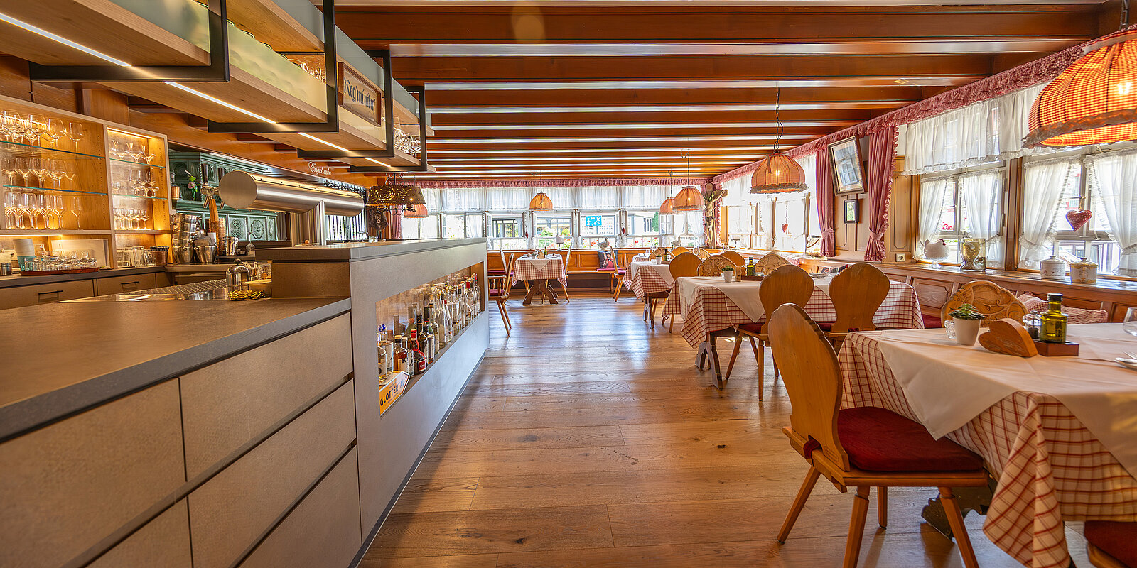 Gemütliches Restaurant mit Holzboden und Holzdecke, eingerichteten Tischen mit karierten Tischdecken und gepolsterten Stühlen. Die Theke auf der linken Seite ist mit Flaschen und Gläsern bestückt. Große Fenster sorgen für viel Tageslicht. Dezente Beleuchtung durch Hängelampen.
