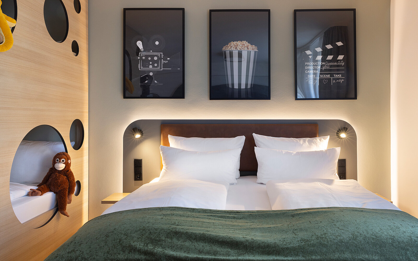 A modern hotel room features a double bed with white pillows and a green throw blanket. Above the bed, three framed movie-themed artworks are displayed. A plush toy monkey is situated on a nearby shelf, adding a whimsical touch to the cozy, stylish décor.
