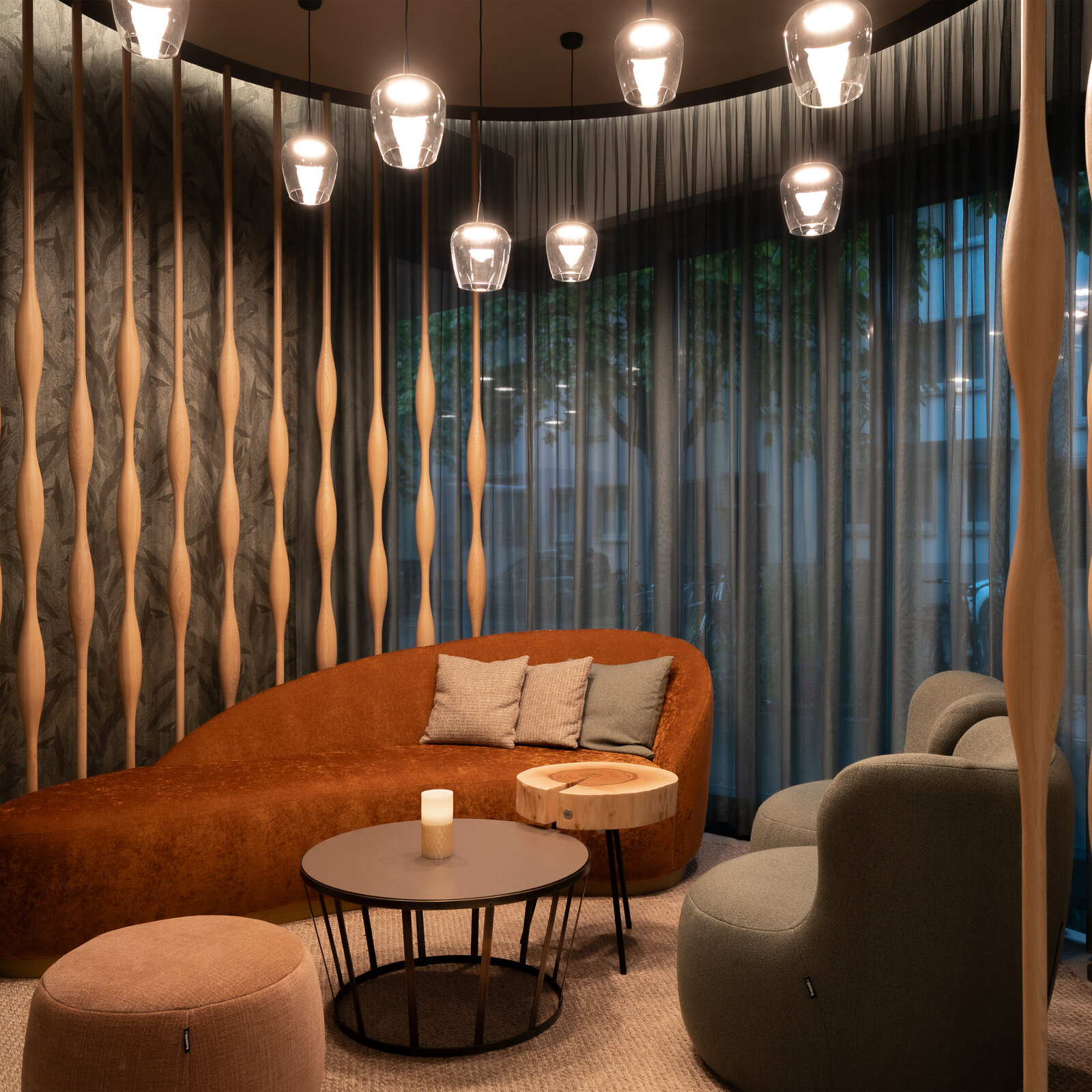 Cozy lounge with a curved brown sofa, round tables, and modern chairs. Soft lighting from hanging lamps and a patterned wall create a warm ambiance.