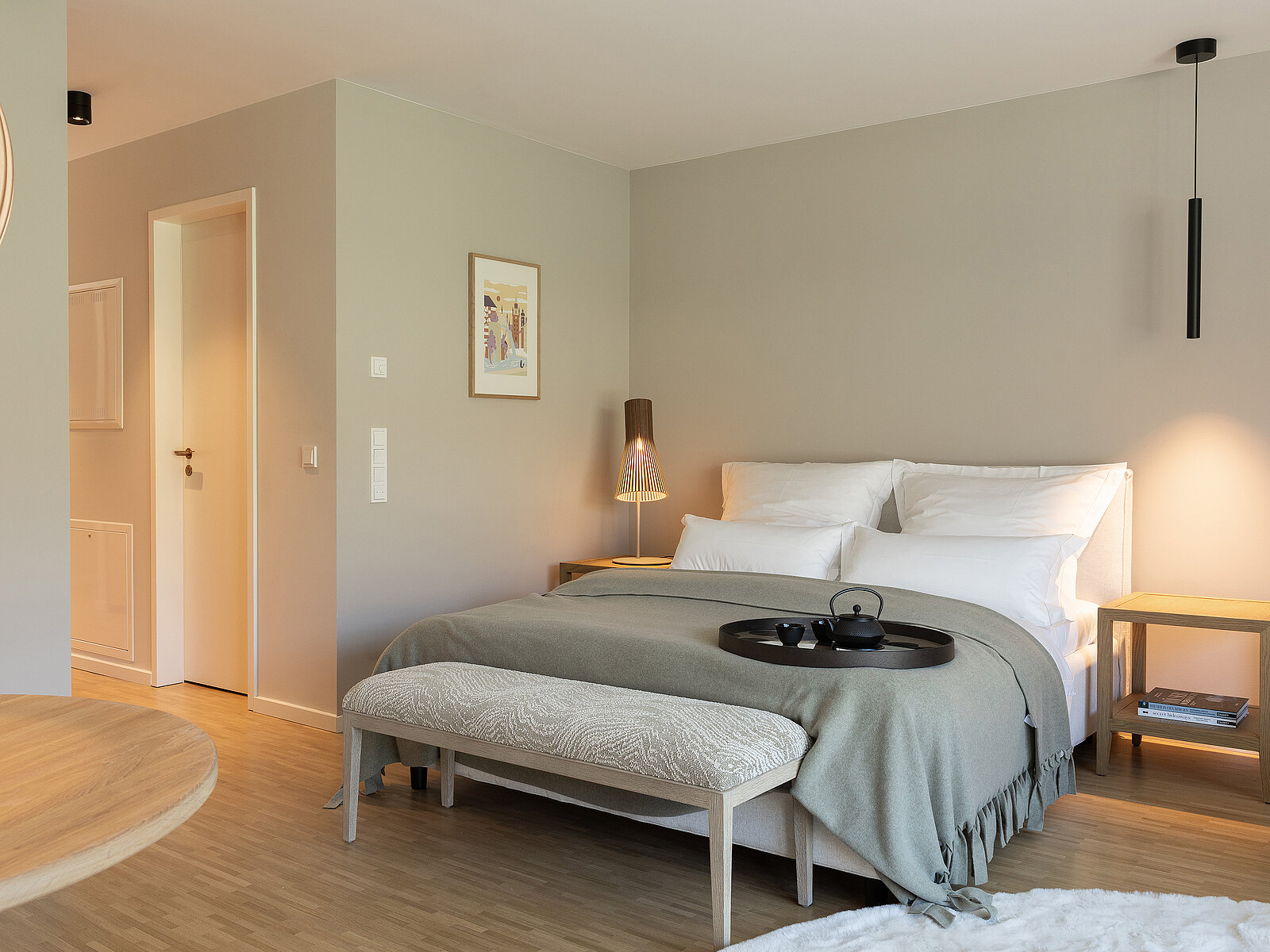 Gemütliches Schlafzimmer mit Doppelbett, hellem Holzfußboden und dezenten Farben. Weiße Bettwäsche und graue Tagesdecke. Nachttische mit Lampe und dekorativem Tablett mit Teekanne auf dem Bett. Schlicht eingerichteter, moderner Raum, der Ruhe ausstrahlt.