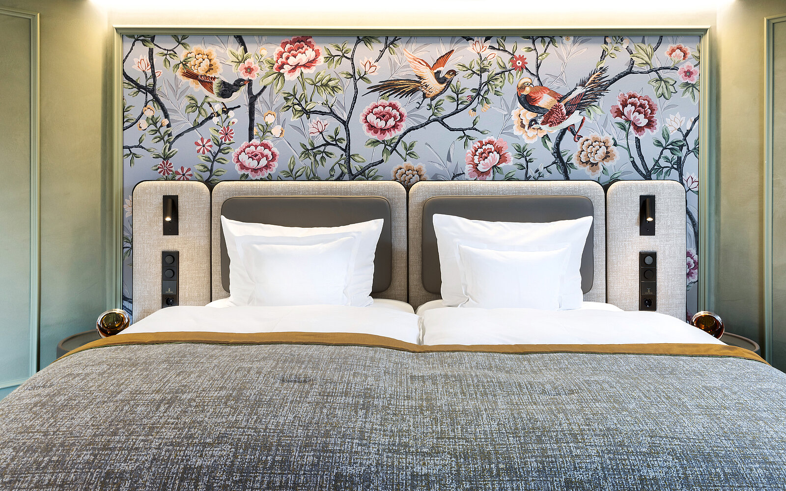 Ein luxuriöses Doppelbett mit eleganten grauen Polstern und weißen Kissen steht vor einer kunstvoll gestalteten Tapete. Die Tapete zeigt ein florales Muster mit Vögeln und Blumen in lebhaften Farben.