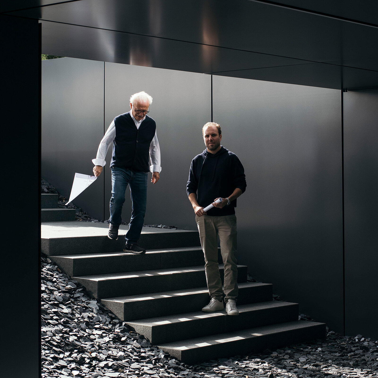 Two men are walking down a set of steps surrounded by dark stones. One man, with white hair, holds a piece of paper and wears a vest, while the other, with shorter hair, holds a phone and wears a dark sweater. They appear engaged in conversation.